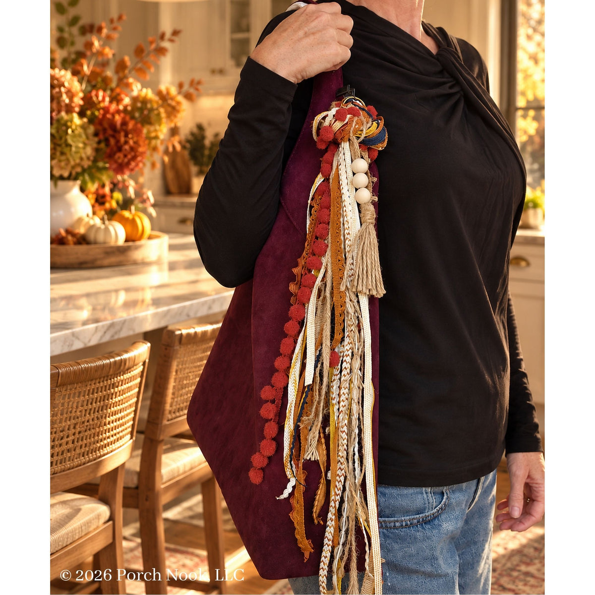 A person wearing a black long-sleeve top and blue jeans carries a large burgundy tote bag decorated with tassels, pom-poms, and woven ribbon in autumn colors, in a warm, sunlit kitchen setting.