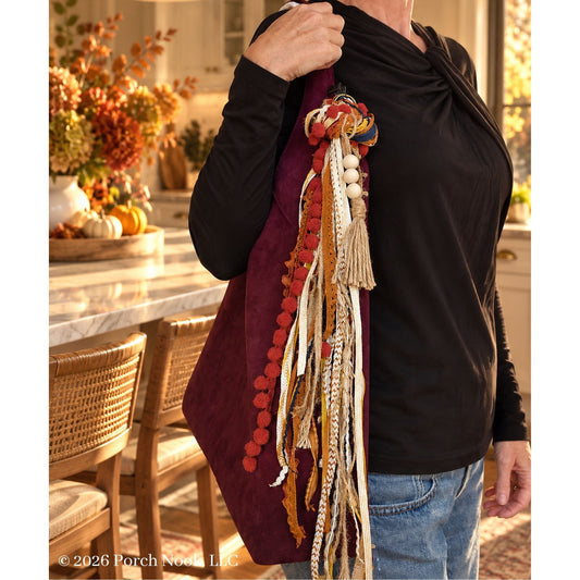 A person wearing a black long-sleeve top and blue jeans carries a large burgundy tote bag decorated with tassels, pom-poms, and woven ribbon in autumn colors, in a warm, sunlit kitchen setting.