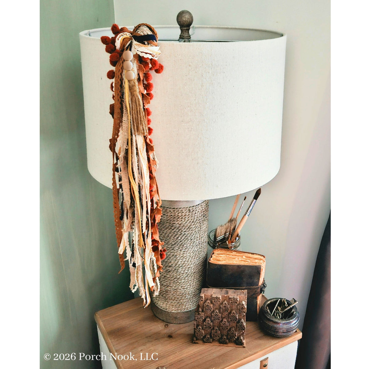 A table lamp with a white drum shade, a wrapped rope base, and a long decorative tassel garland in rust orange, beige, and brown hanging from the lamp. The lamp sits on a wooden side table against a pale green wall, with small decor items beside it.