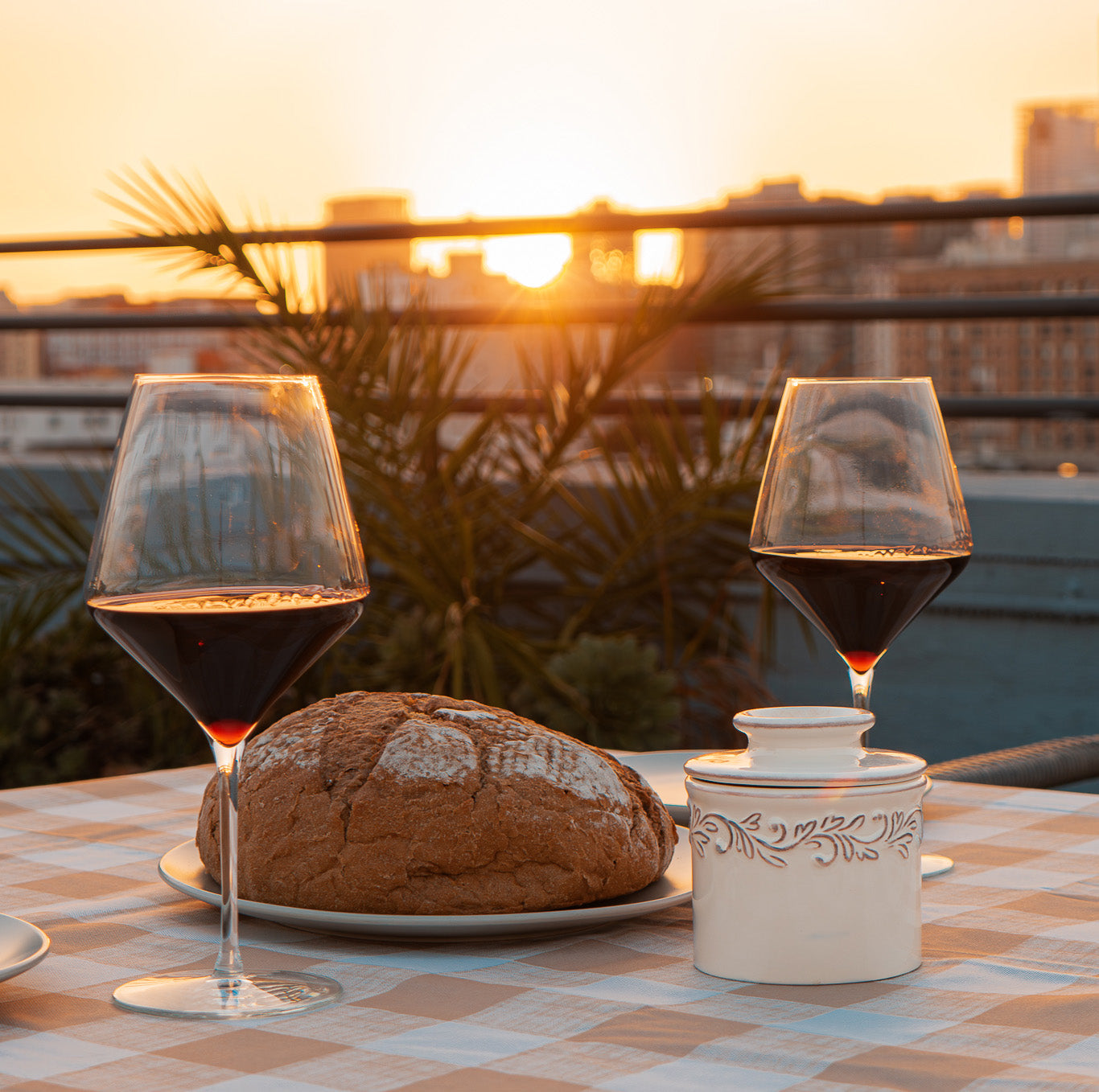 Two glasses of red wine, a loaf of bread, and a small container on a table with a sunset cityscape in the background.