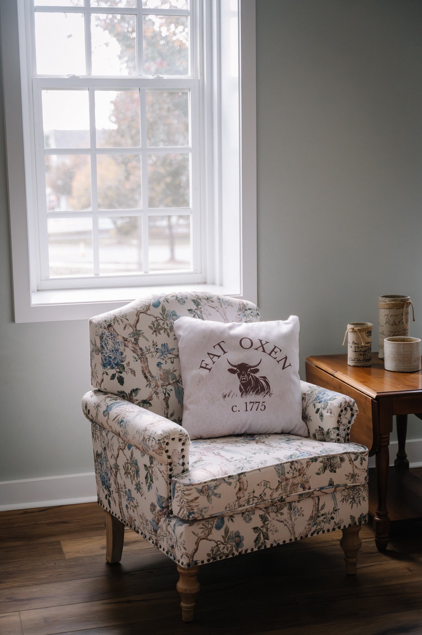 Floral-patterned armchair with a 'Fat Oxen' pillow in a room with a window and wooden side table.
