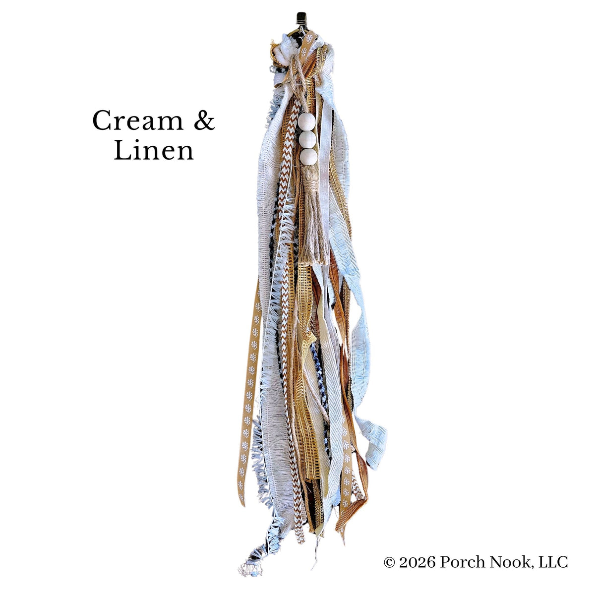 A decorative hanging textile piece labeled 'Cream & Linen' on a white background. The piece features layered ribbons, fringe, tassels, and woven details in cream, beige, tan, and light blue, with a small black top and white circular accents near the top.