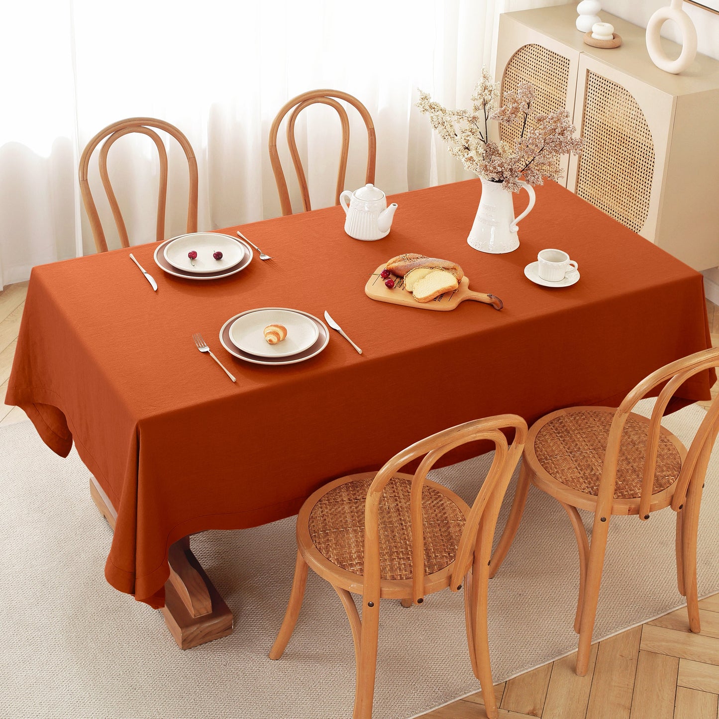 An orange-brown rectangular tablecloth draped over a dining table set with wooden chairs, plates, cutlery, a teapot, and a vase of dried flowers in a bright room.