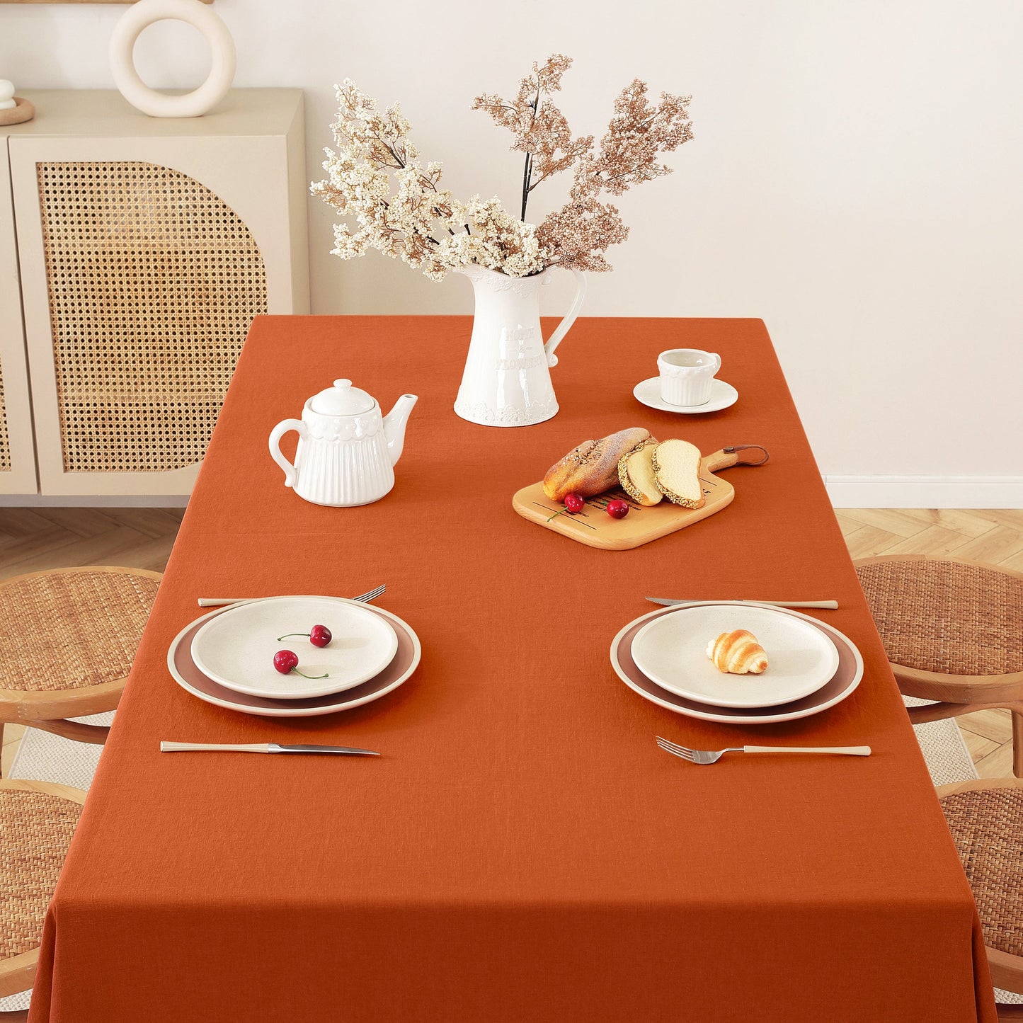 An orange-brown rectangular tablecloth covers a dining table set with white plates, a white teapot, a teacup and saucer, a small cutting board with bread and berries, and a vase of dried flowers. Chairs and a cabinet are visible in a bright indoor room.