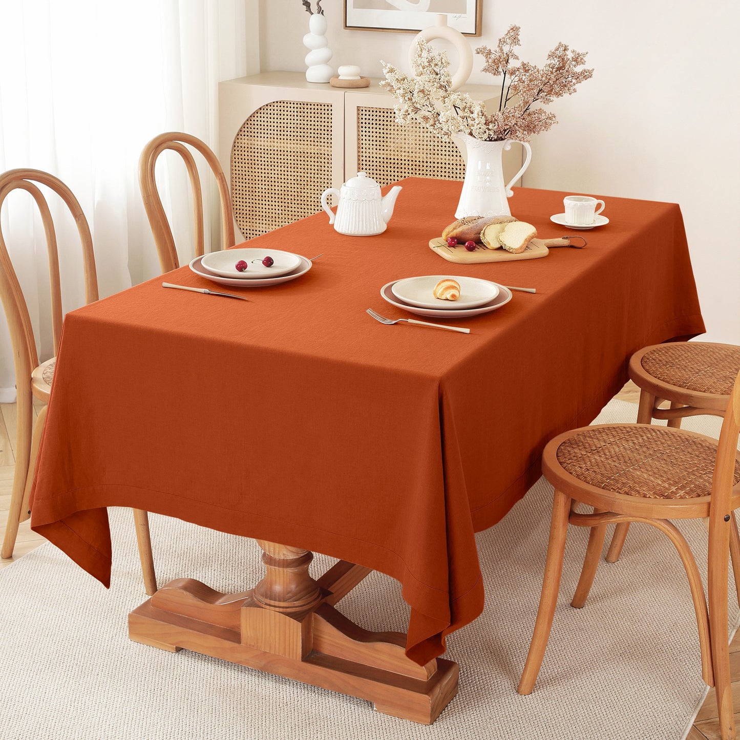 A rust-orange rectangular tablecloth draped over a wooden dining table set with white plates, a white teapot, and a vase of dried flowers in a bright, neutral-toned dining room.