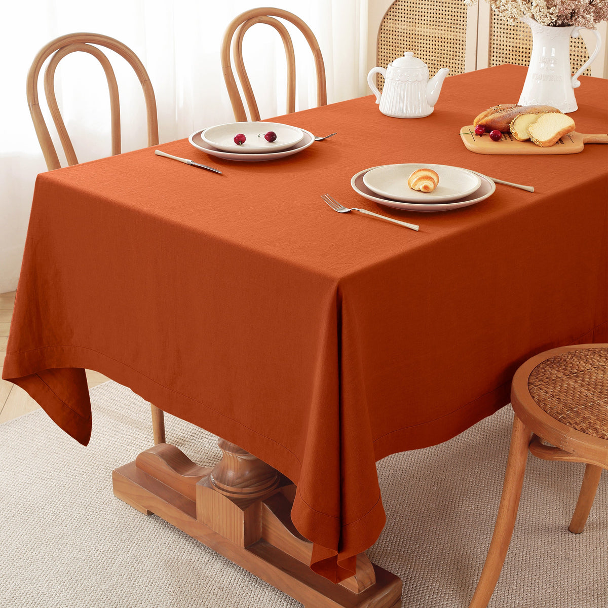 A rust-orange rectangular tablecloth draped over a wooden dining table, styled with white plates, silver cutlery, a white teapot, and pastries on a light beige carpeted floor with wooden chairs.