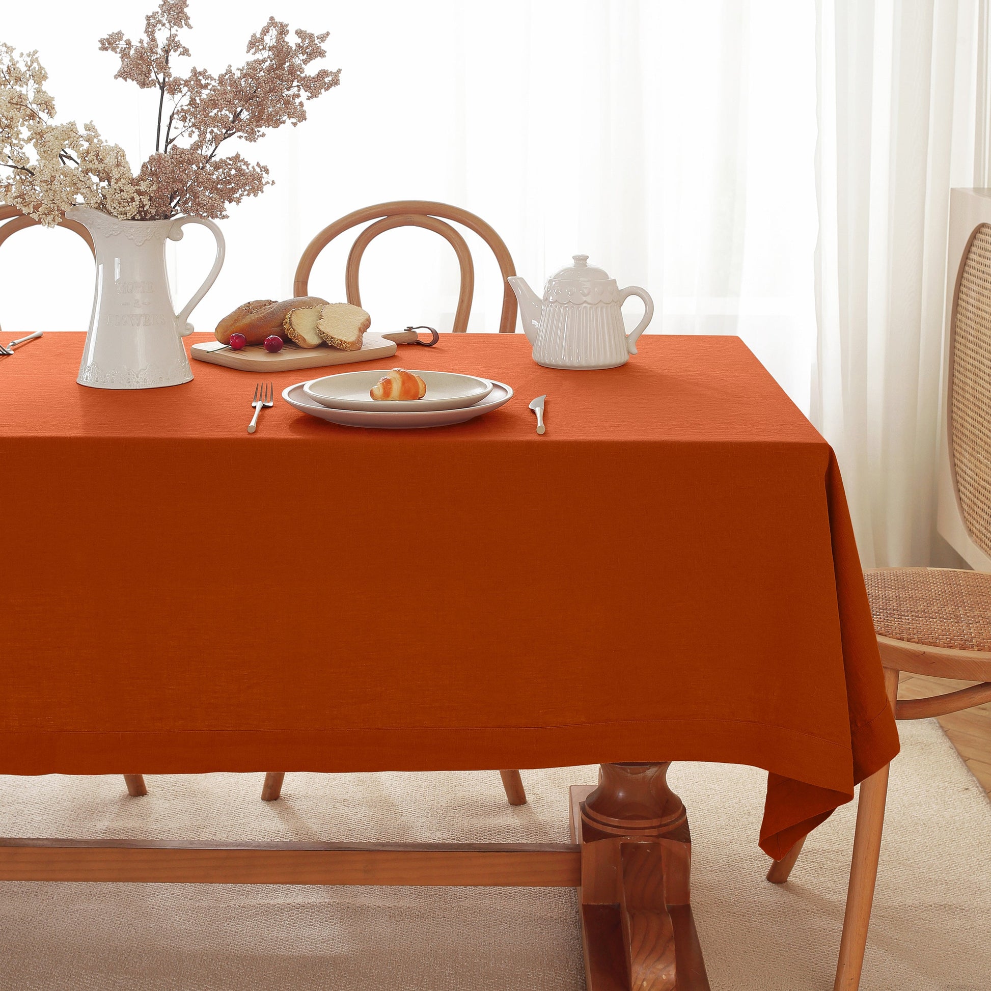 An orange rectangular tablecloth draped over a wooden dining table, styled with a white teapot, plates, bread, and a white pitcher with dried flowers in a bright dining room.