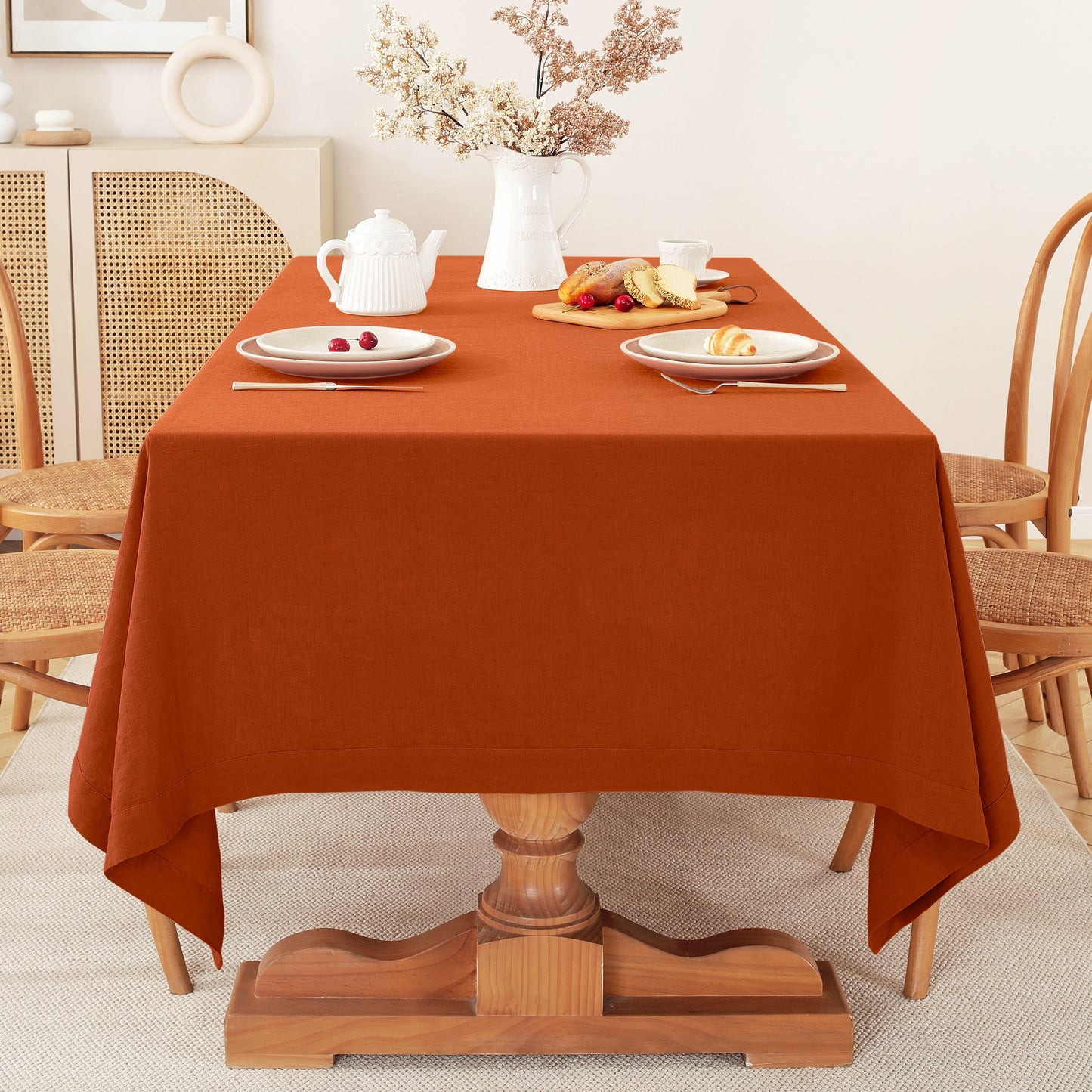An orange-brown rectangular tablecloth draped over a wooden dining table set with white plates, a white teapot, and a vase of dried flowers in a warm, neutral-toned dining room.