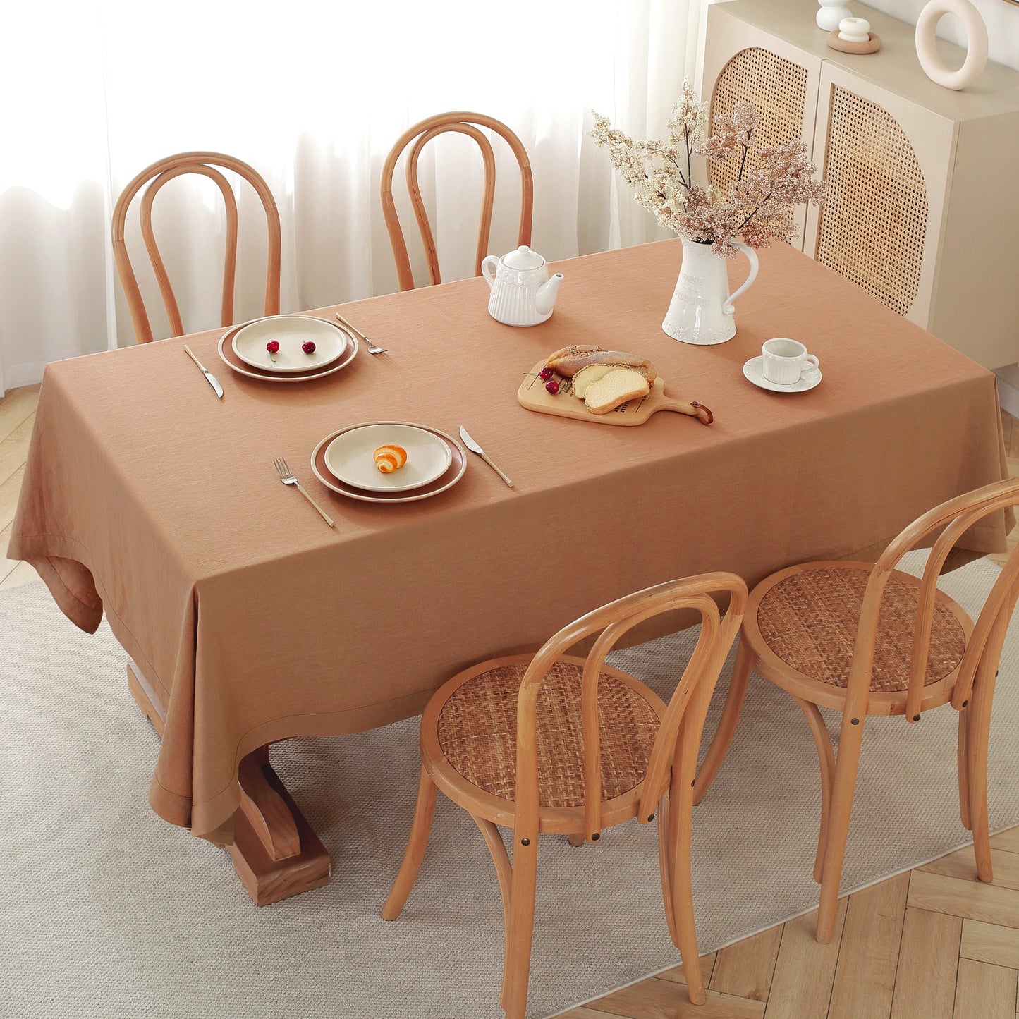 A rectangular dining table covered with a warm tan-brown tablecloth, set with plates, cutlery, a white teapot, a vase of dried flowers, and a small bread dish. Four wooden chairs with woven seats surround the table in a bright, softly lit room.
