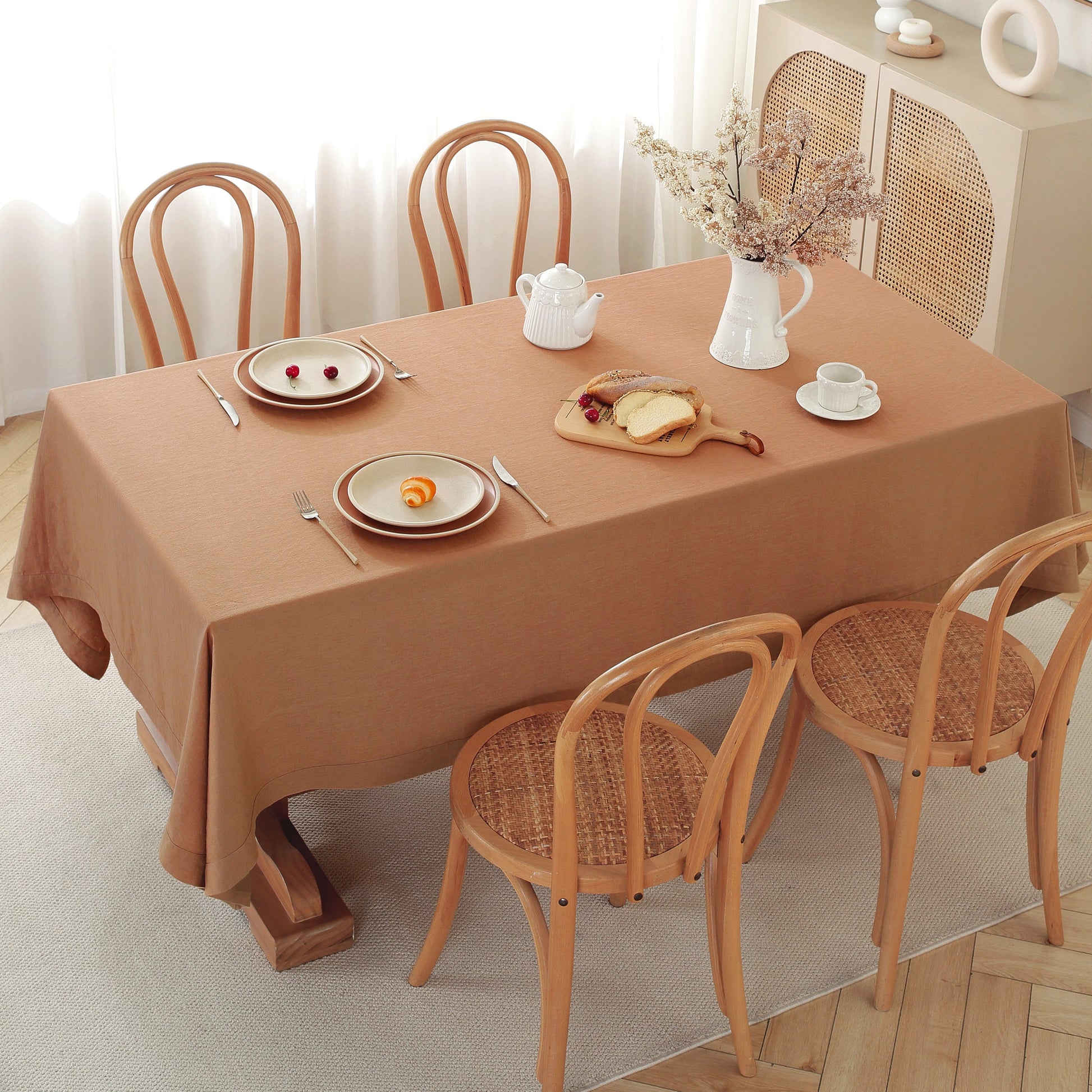 A rectangular dining table covered with a warm tan-brown tablecloth, set with plates, cutlery, a white teapot, a vase of dried flowers, and a small bread dish. Four wooden chairs with woven seats surround the table in a bright, softly lit room.