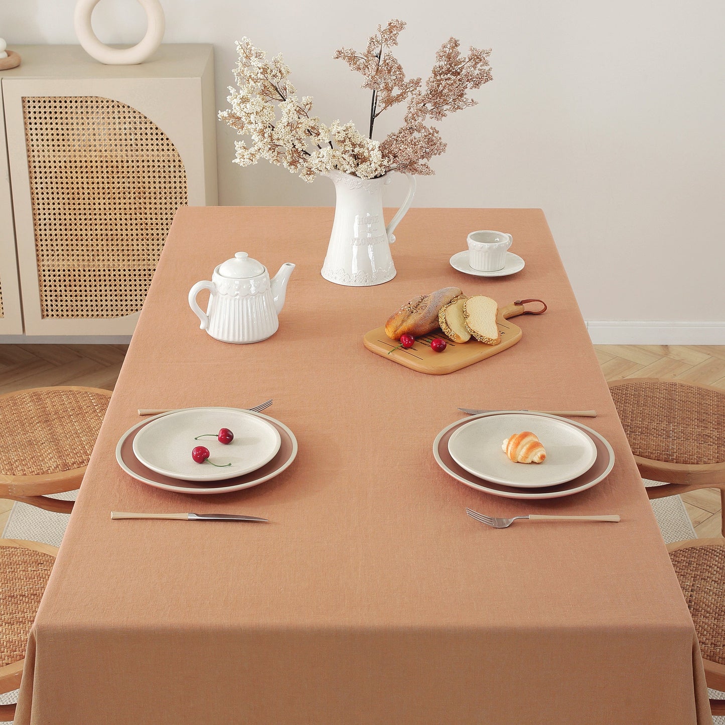 A rectangular dining table covered with a warm beige tablecloth, styled with two place settings, a white teapot, a vase of dried flowers, and a small bread board. The scene is photographed in a bright, neutral room with woven chairs and a cabinet.