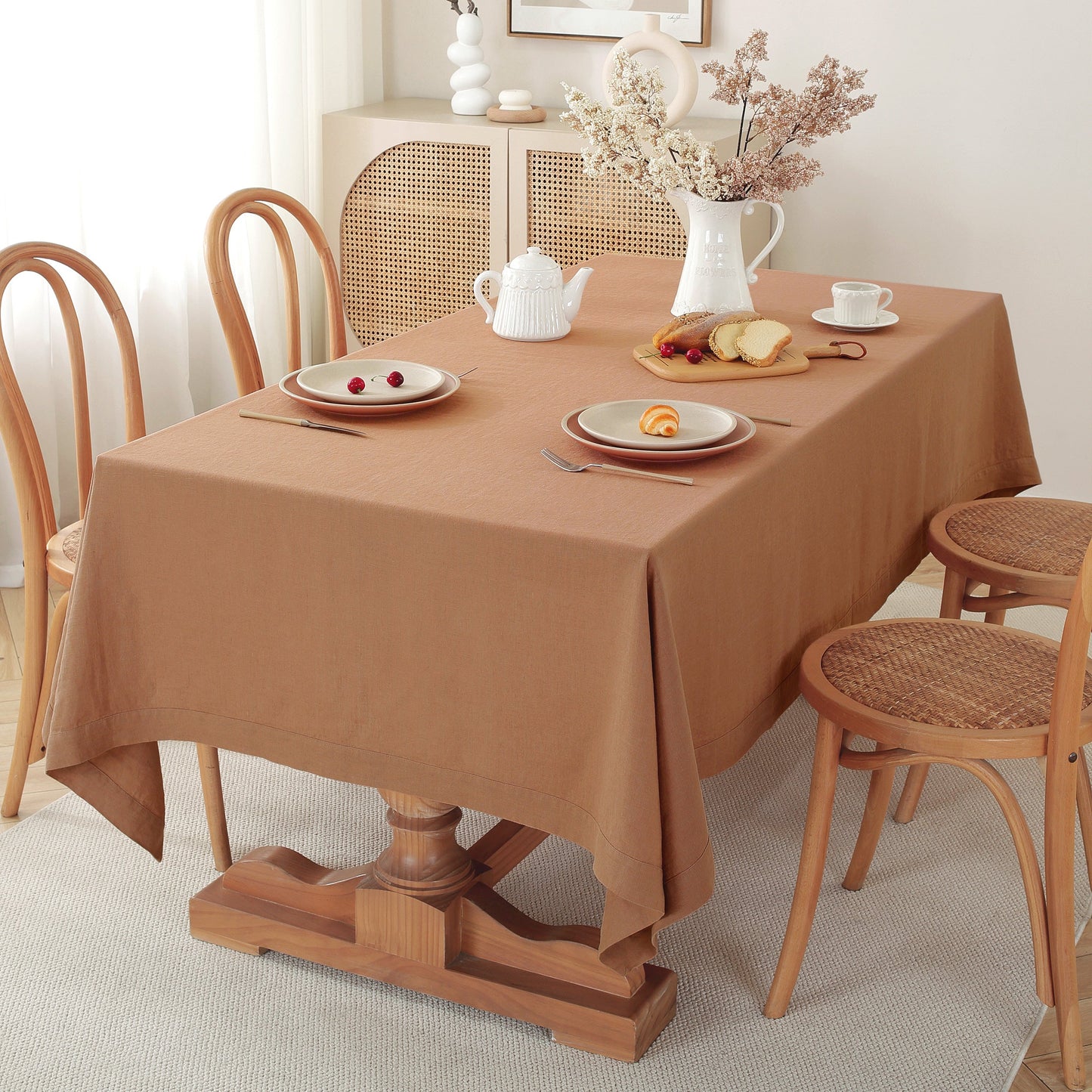 A warm brown tablecloth draped over a wooden dining table set with plates, a white teapot, and a vase of dried flowers in a softly lit room.