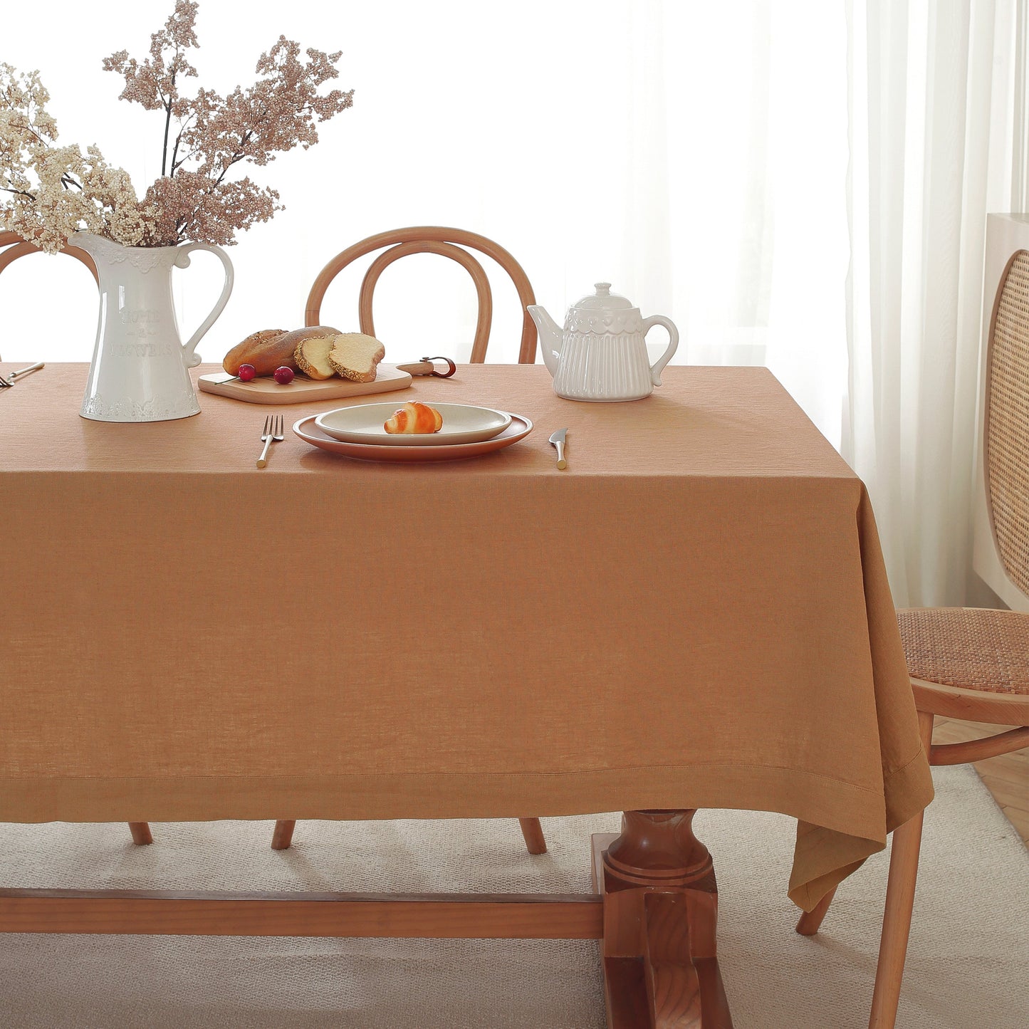 A warm tan tablecloth draped over a wooden dining table, styled with a white pitcher of dried flowers, bread, plates, and a white teapot in a bright, softly lit room.