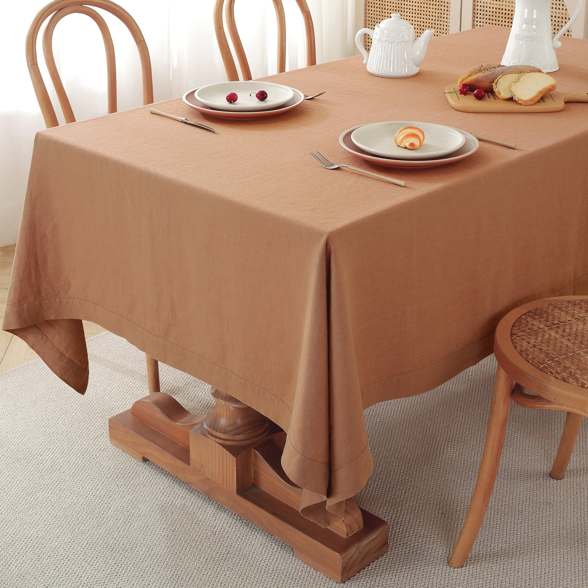 A warm brown tablecloth draped over a wooden dining table, styled with white plates, silver cutlery, a white teapot, and a small bread board, with wooden chairs in a bright, neutral room.