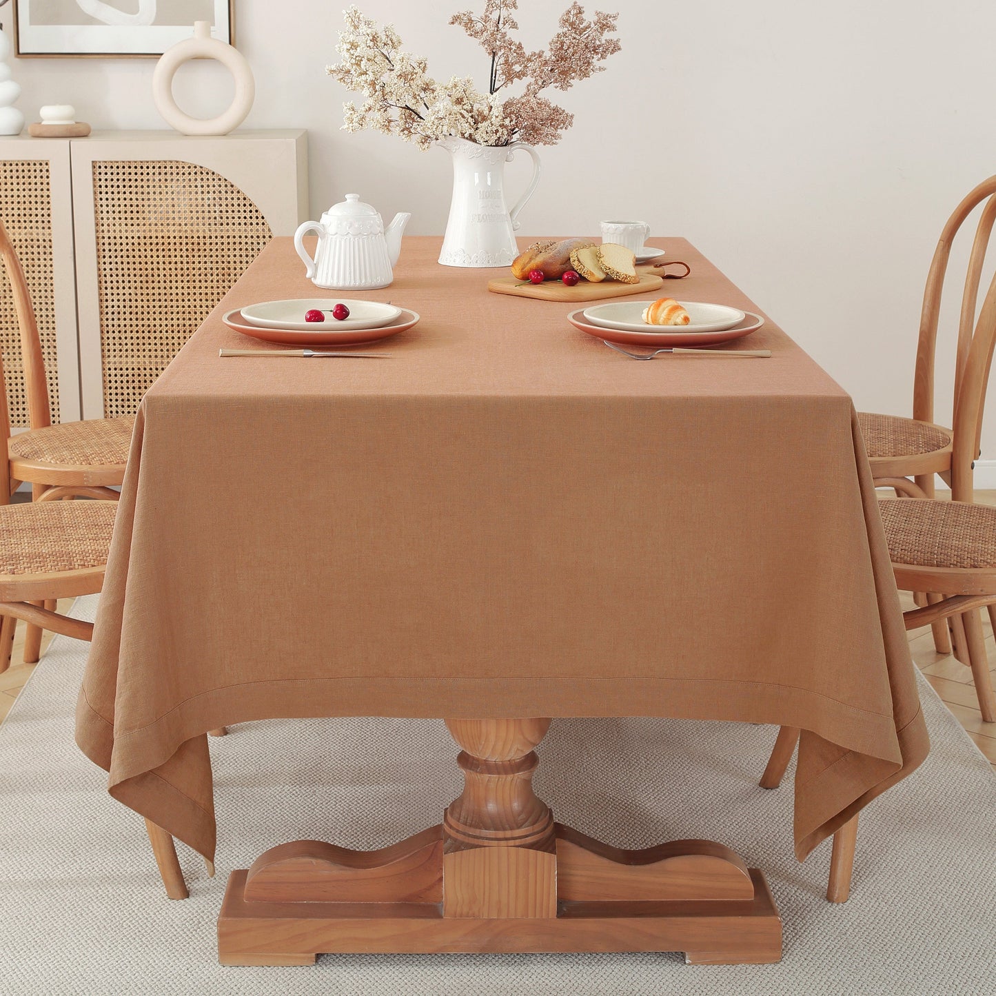 A warm brown tablecloth draped over a wooden dining table in a bright, neutral-toned dining room setting with chairs, a teapot, and plates.