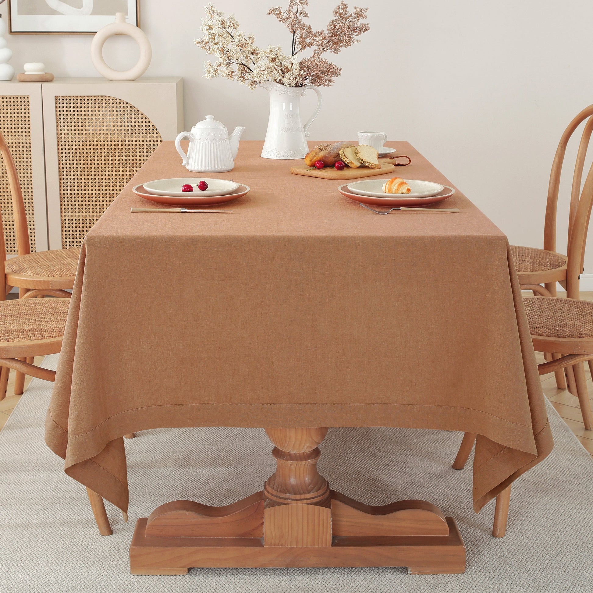 A warm brown tablecloth draped over a wooden dining table in a bright, neutral-toned dining room setting with chairs, a teapot, and plates.