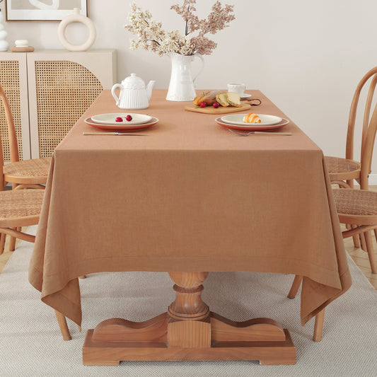 A warm brown tablecloth draped over a wooden dining table in a bright, neutral-toned dining room setting with chairs, a teapot, and plates.
