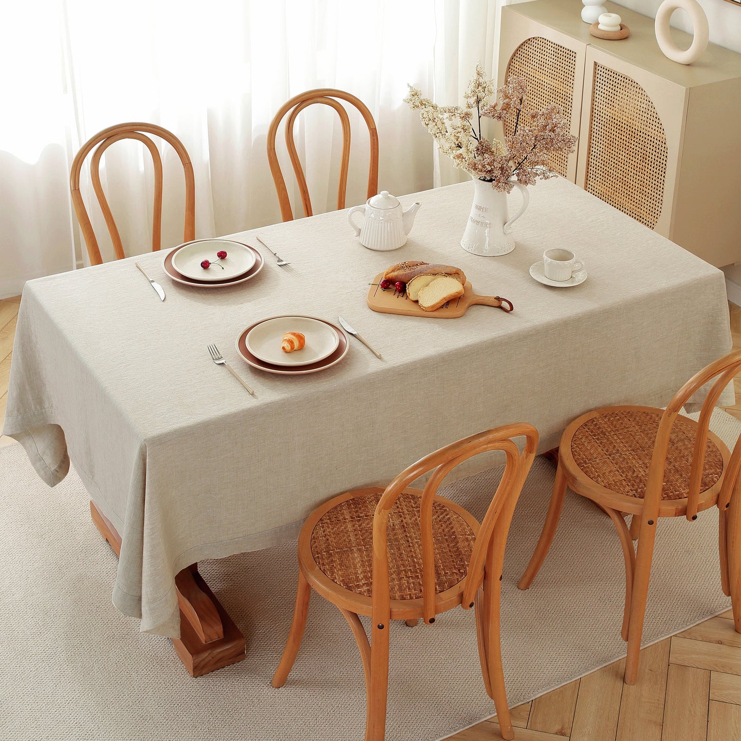 A rectangular dining table covered with a light beige tablecloth, surrounded by four wooden chairs with woven seats. The table is set with plates, cutlery, a white teapot, and a vase of dried flowers, in a bright, softly lit room.