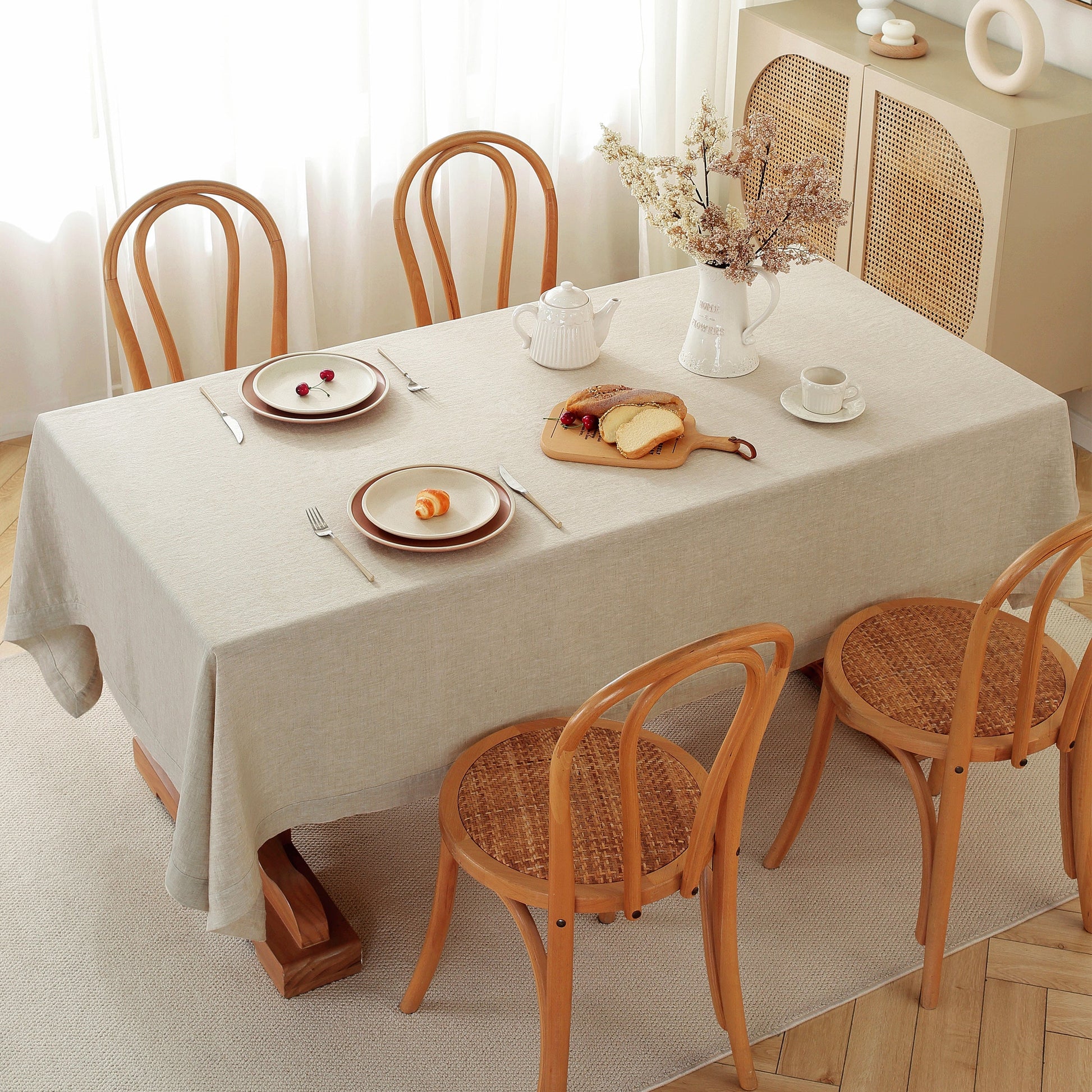 A rectangular dining table covered with a light beige tablecloth, surrounded by four wooden chairs with woven seats. The table is set with plates, cutlery, a white teapot, and a vase of dried flowers, in a bright, softly lit room.