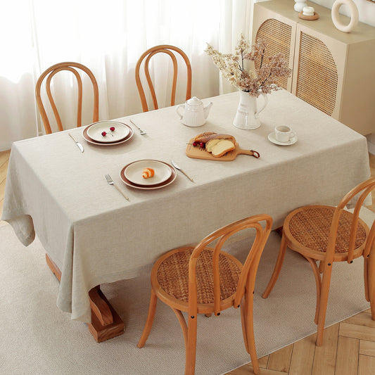 A rectangular dining table covered with a light beige tablecloth, surrounded by four wooden chairs with woven seats. The table is set with plates, cutlery, a white teapot, and a vase of dried flowers, in a bright, softly lit room.