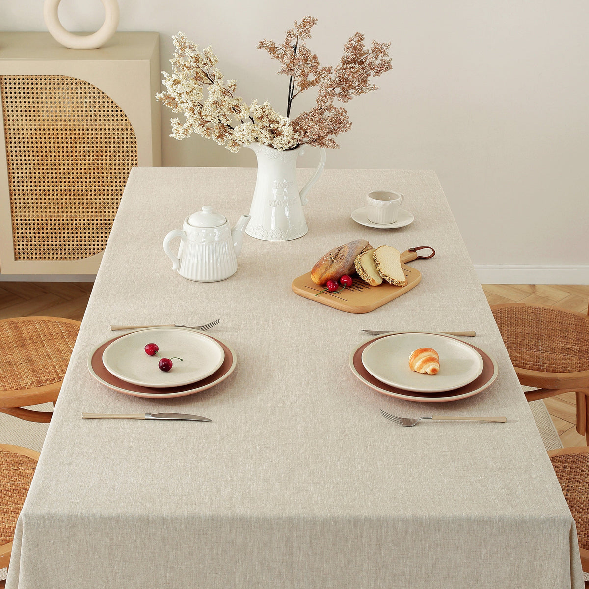 A light beige linen tablecloth draped over a rectangular dining table, styled with white ceramic dishes, a teapot, a vase of dried flowers, and a small wooden board with bread and cherries. The scene is set in a warm, neutral-toned room with woven rattan chairs and a matching cabinet.
