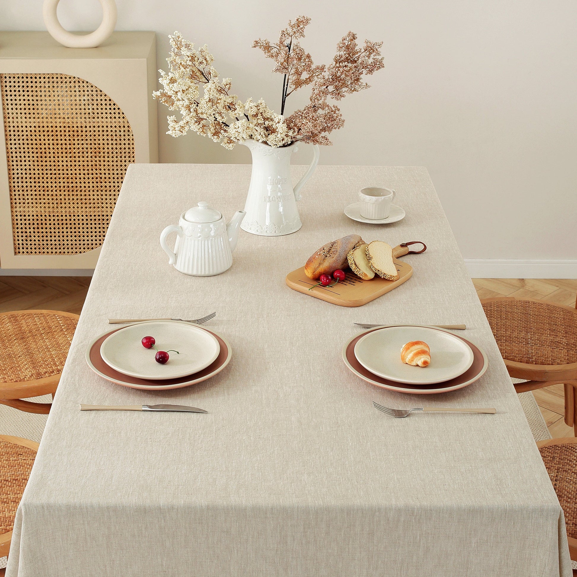 A light beige linen tablecloth draped over a rectangular dining table, styled with white ceramic dishes, a teapot, a vase of dried flowers, and a small wooden board with bread and cherries. The setting includes woven rattan chairs and a matching sideboard against a warm cream wall.