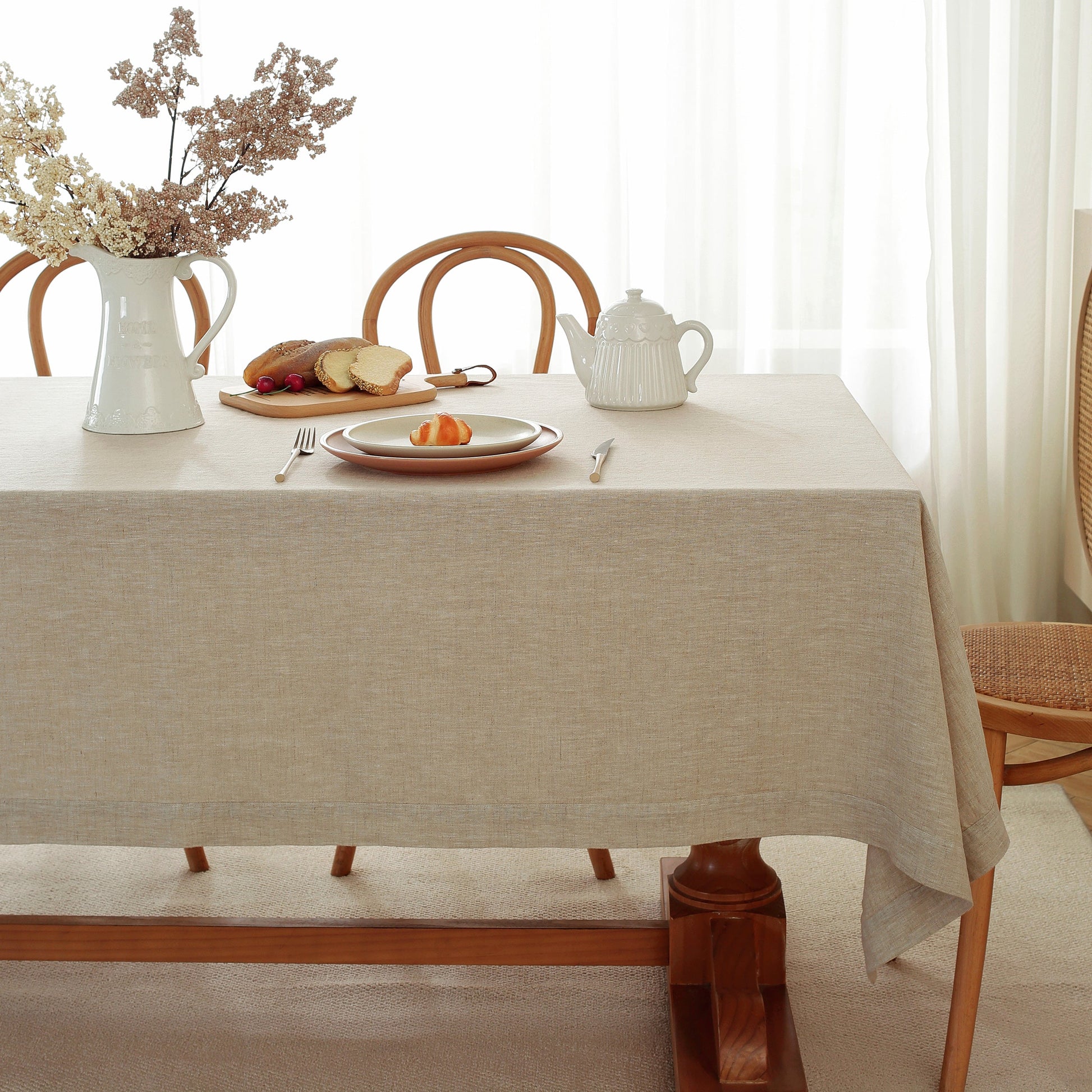 A light beige linen tablecloth draped over a wooden dining table, styled with a white pitcher of dried flowers, a white teapot, and plates with bread on a wooden chair and table in a bright, softly lit room.