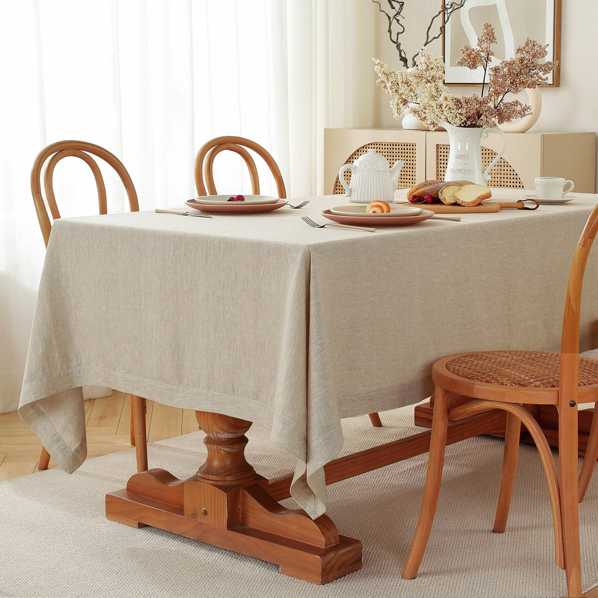 A light beige linen tablecloth draped over a wooden dining table set with plates, cutlery, and chairs in a bright, softly lit room.
