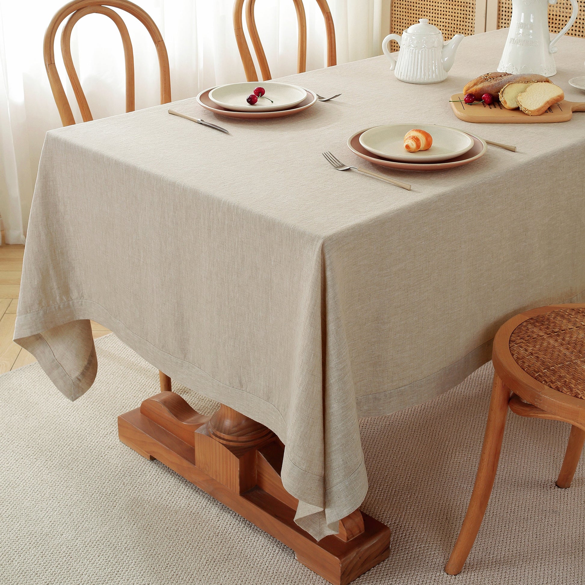 A light beige linen tablecloth draped over a wooden dining table set with plates, cutlery, a white teapot, and bread, in a bright, softly lit room.