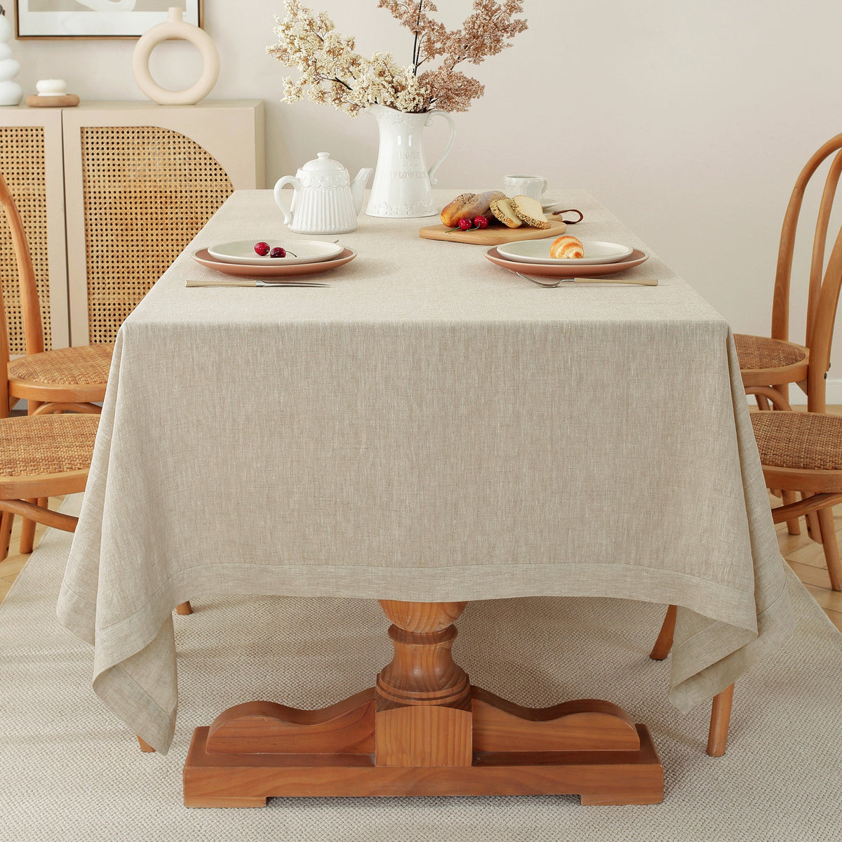 A light beige linen tablecloth draped over a wooden pedestal dining table, styled with plates, a white teapot, and dried flowers in the background.