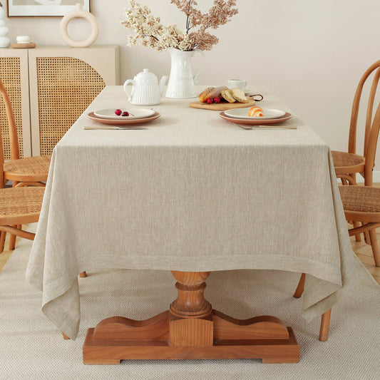 A light beige linen tablecloth draped over a wooden pedestal dining table, styled with plates, a white teapot, and dried flowers in the background.