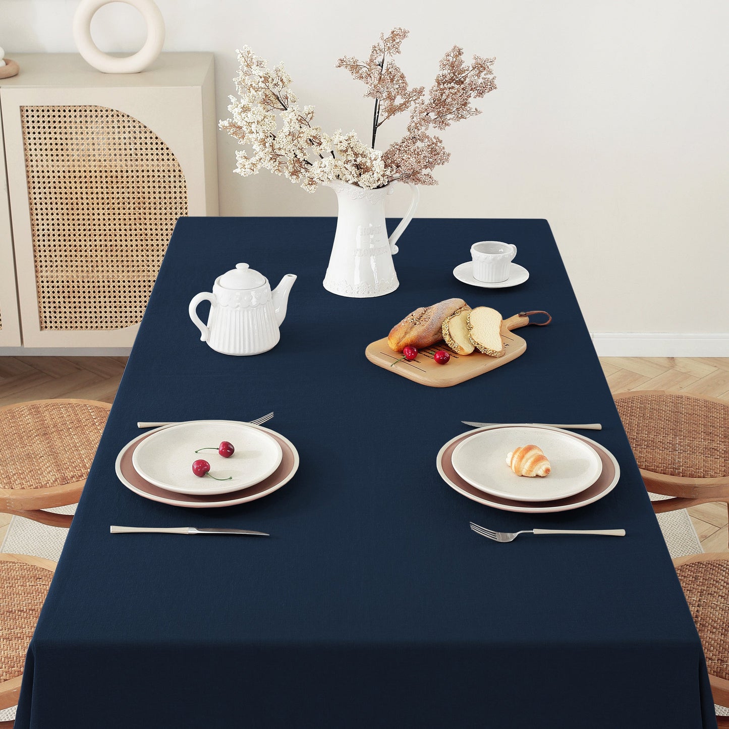 A navy blue tablecloth draped over a rectangular dining table, styled with white plates, silver cutlery, a white teapot, a cup and saucer, bread on a wooden board, and a vase of dried flowers in a bright room.