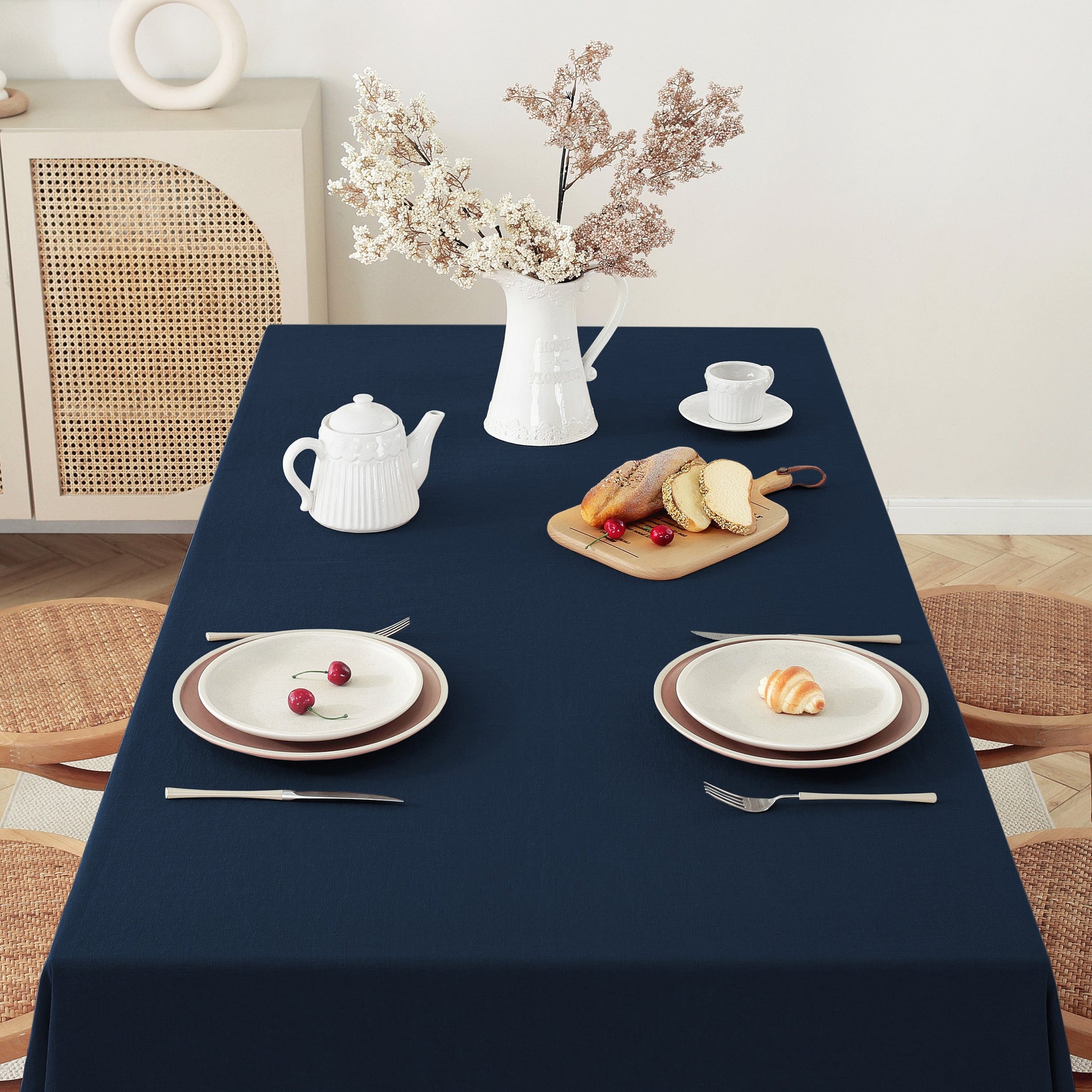 A navy blue tablecloth draped over a rectangular dining table, styled with white plates, silver cutlery, a white teapot, a cup and saucer, bread on a wooden board, and a vase of dried flowers in a bright room.