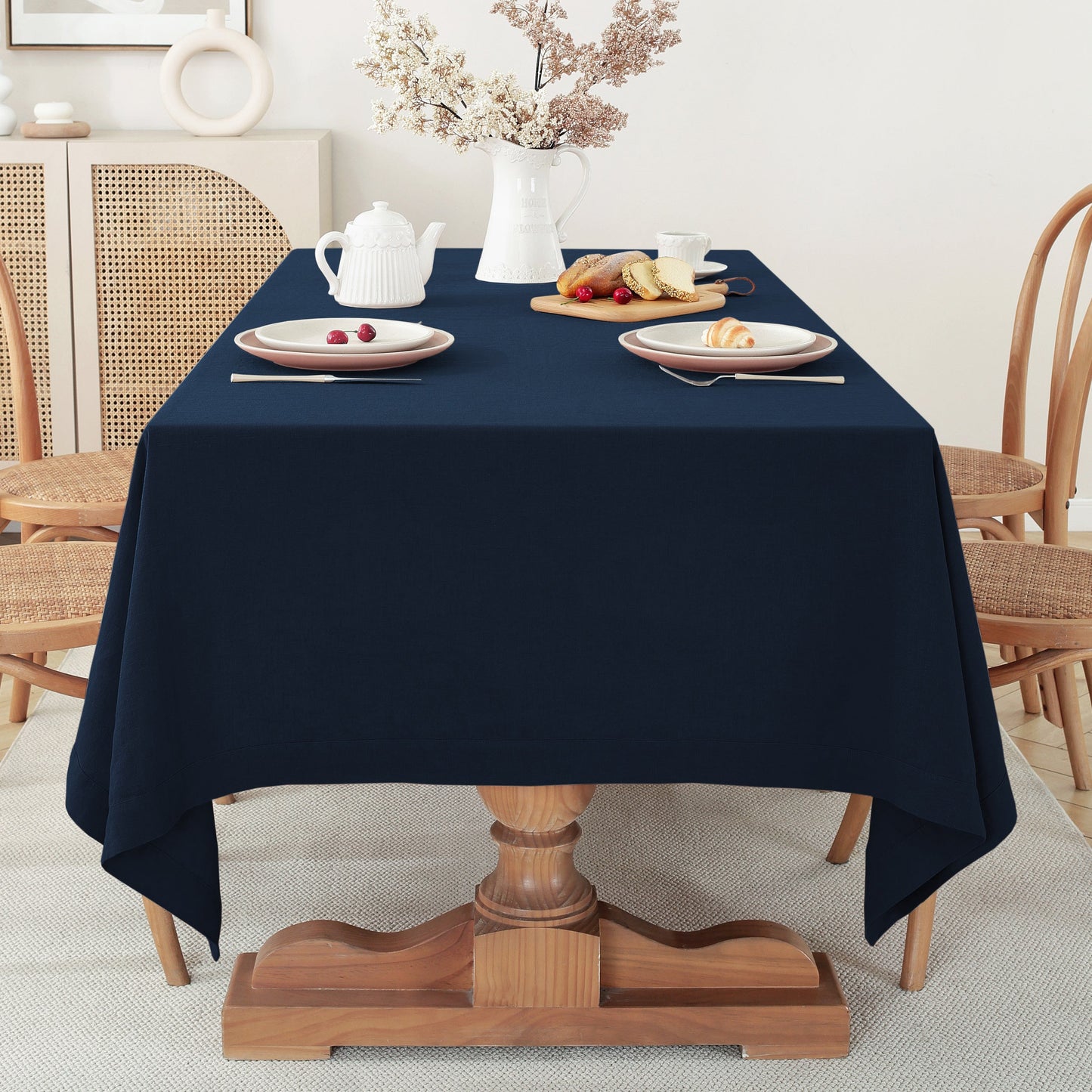 A navy blue tablecloth draped over a wooden pedestal dining table, styled with plates, a white teapot, and a vase of dried flowers in a bright, neutral room.