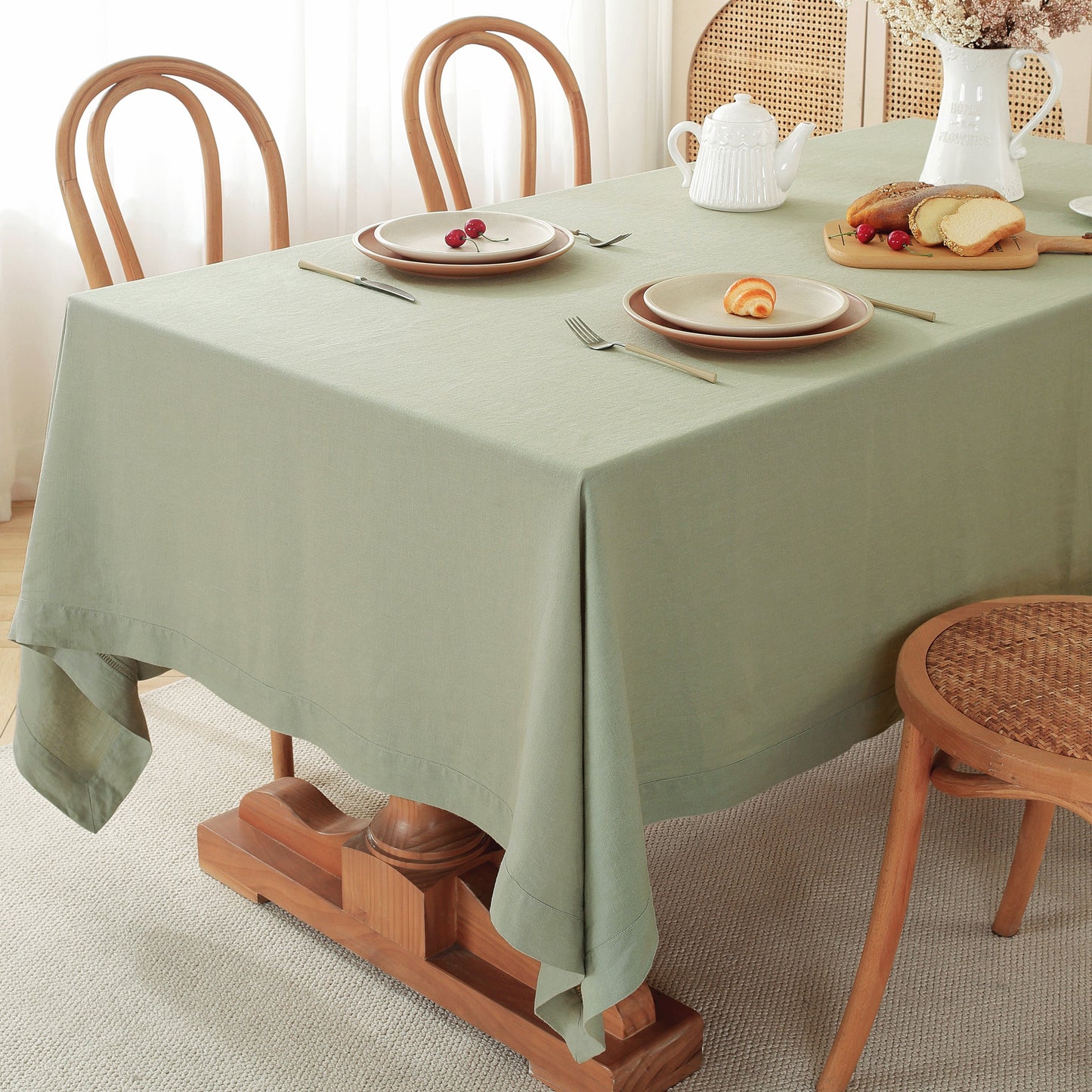 A sage green tablecloth draped over a wooden dining table set with plates, cutlery, a teapot, and bread, in a bright, neutral room with wooden chairs.