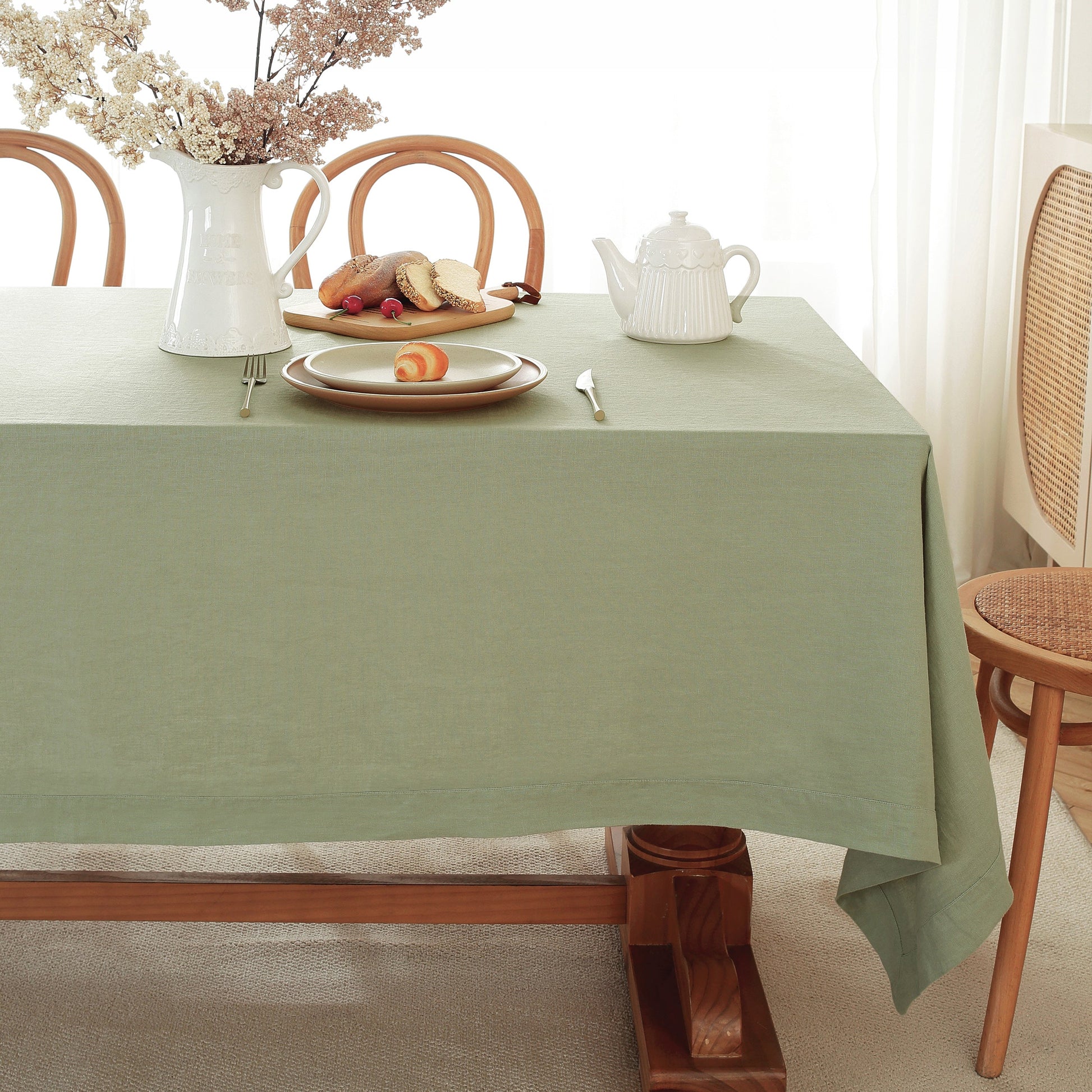 A sage green tablecloth draped over a wooden dining table, styled with a white ceramic pitcher of flowers, a plate with bread and fruit, a white teapot, and silver cutlery. Wooden dining chairs are visible in a bright, softly lit room.