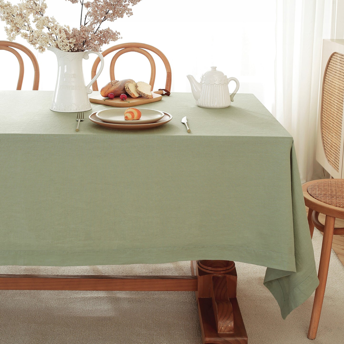 A sage green tablecloth draped over a wooden dining table, styled with a white ceramic pitcher of flowers, a plate with bread and fruit, a white teapot, and silver cutlery. Wooden dining chairs are visible in a bright, softly lit room.