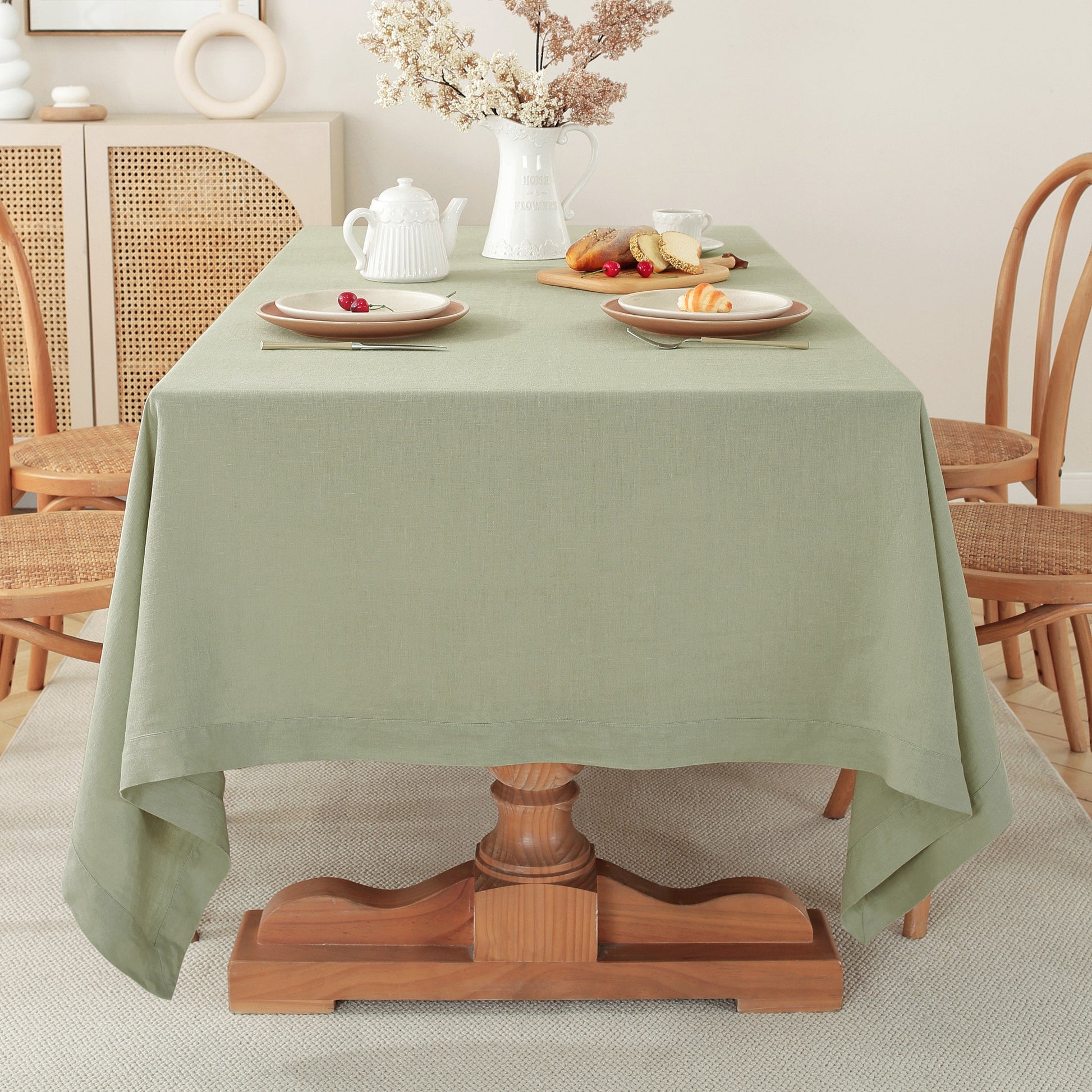 A sage green tablecloth draped over a wooden pedestal dining table, styled with plates, a white teapot, and a vase of dried flowers in a warm, neutral-toned dining room.