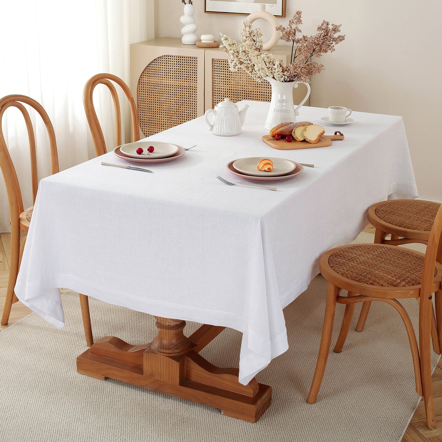 A white tablecloth draped over a wooden dining table set with plates, cutlery, a teapot, and a vase of dried flowers, surrounded by wooden chairs in a bright dining room.