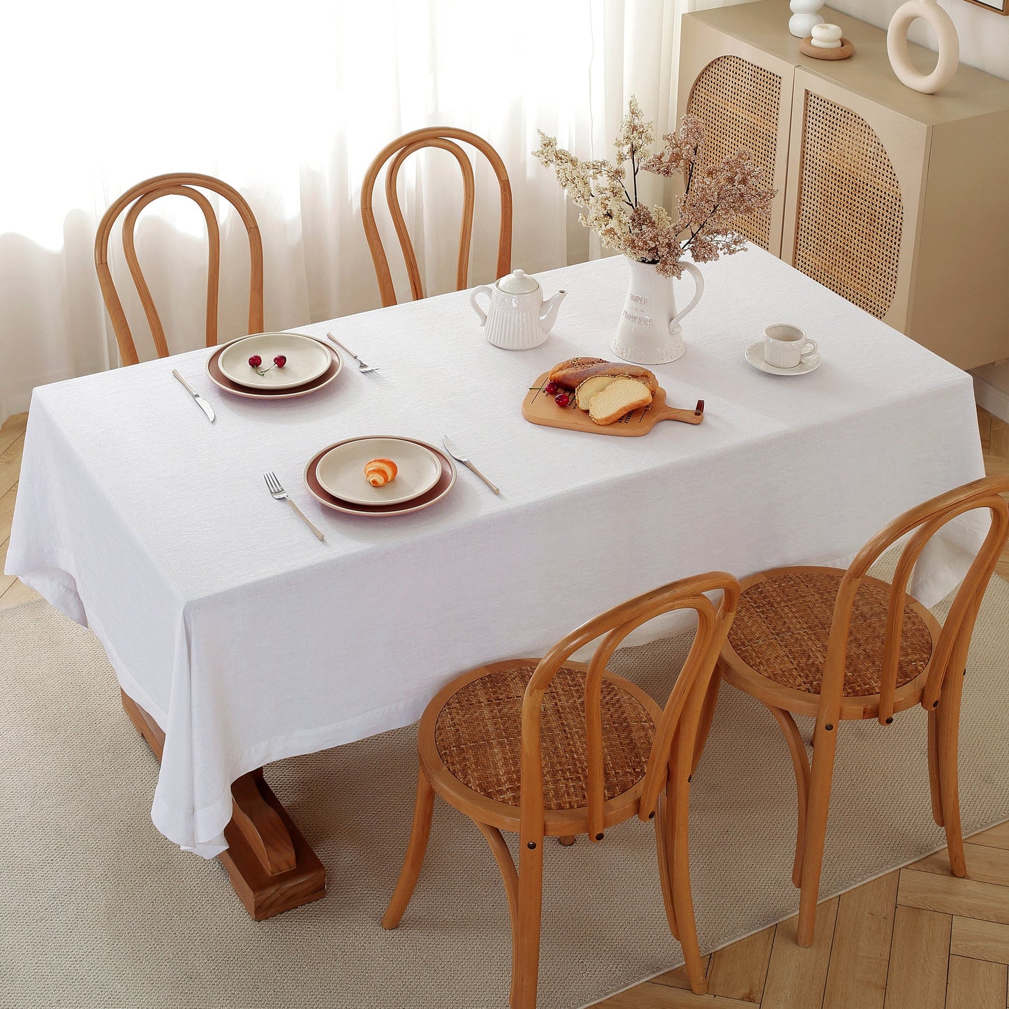 Dining table set with plates, teapot, and bread on a white tablecloth with wooden chairs.