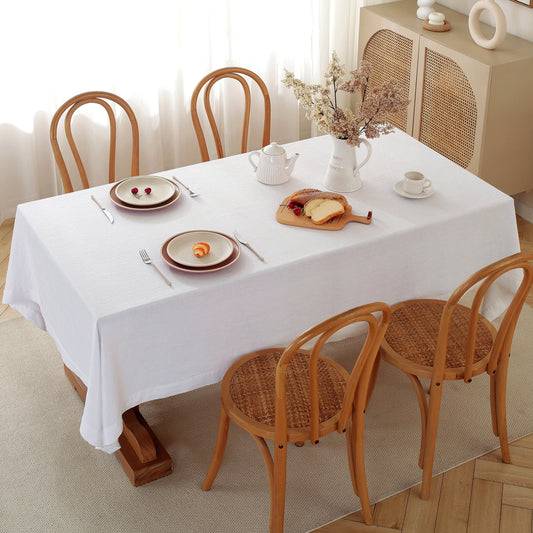 Dining table set with plates, teapot, and bread on a white tablecloth with wooden chairs.