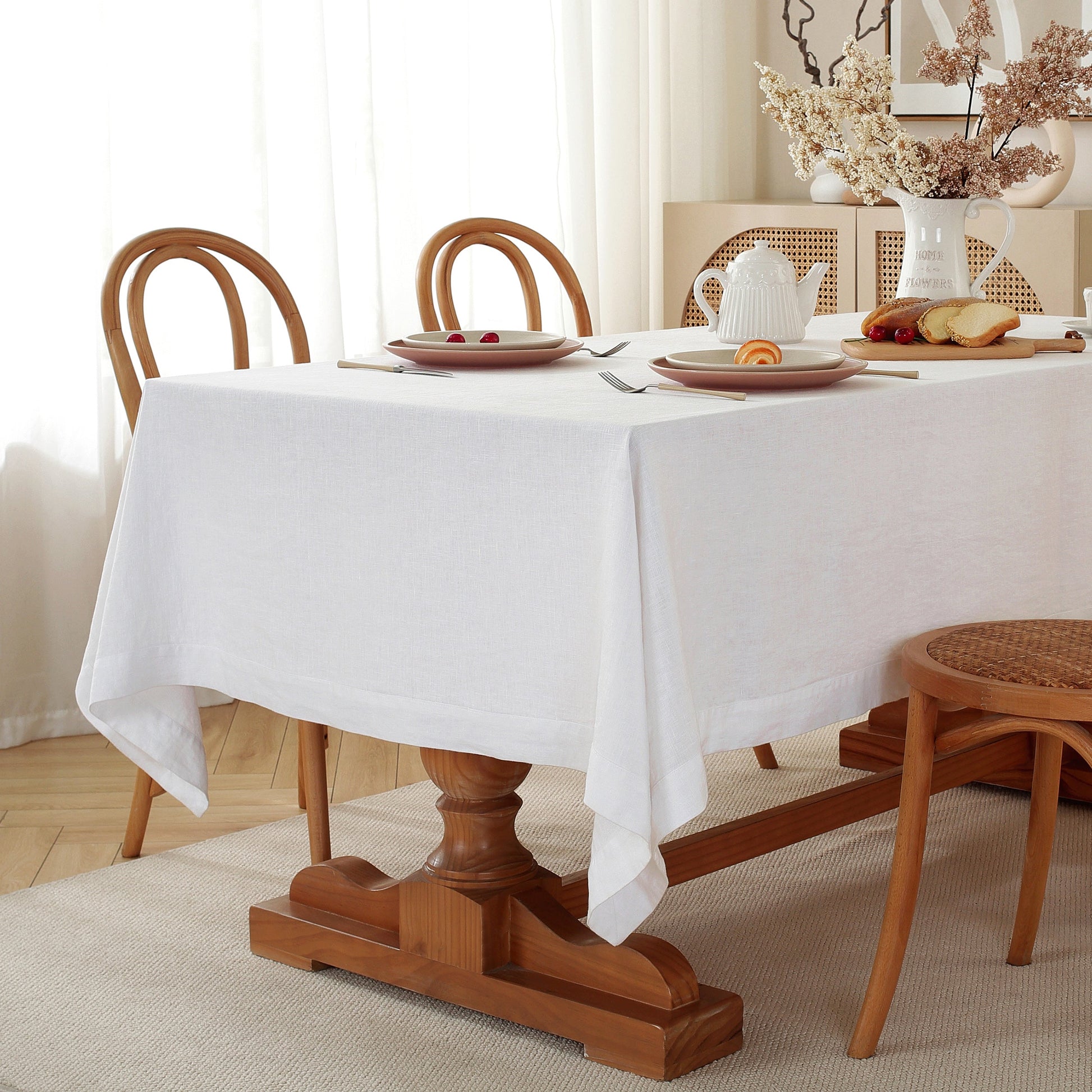 A white tablecloth draped over a wooden dining table in a bright, softly lit room with wooden chairs and tableware.