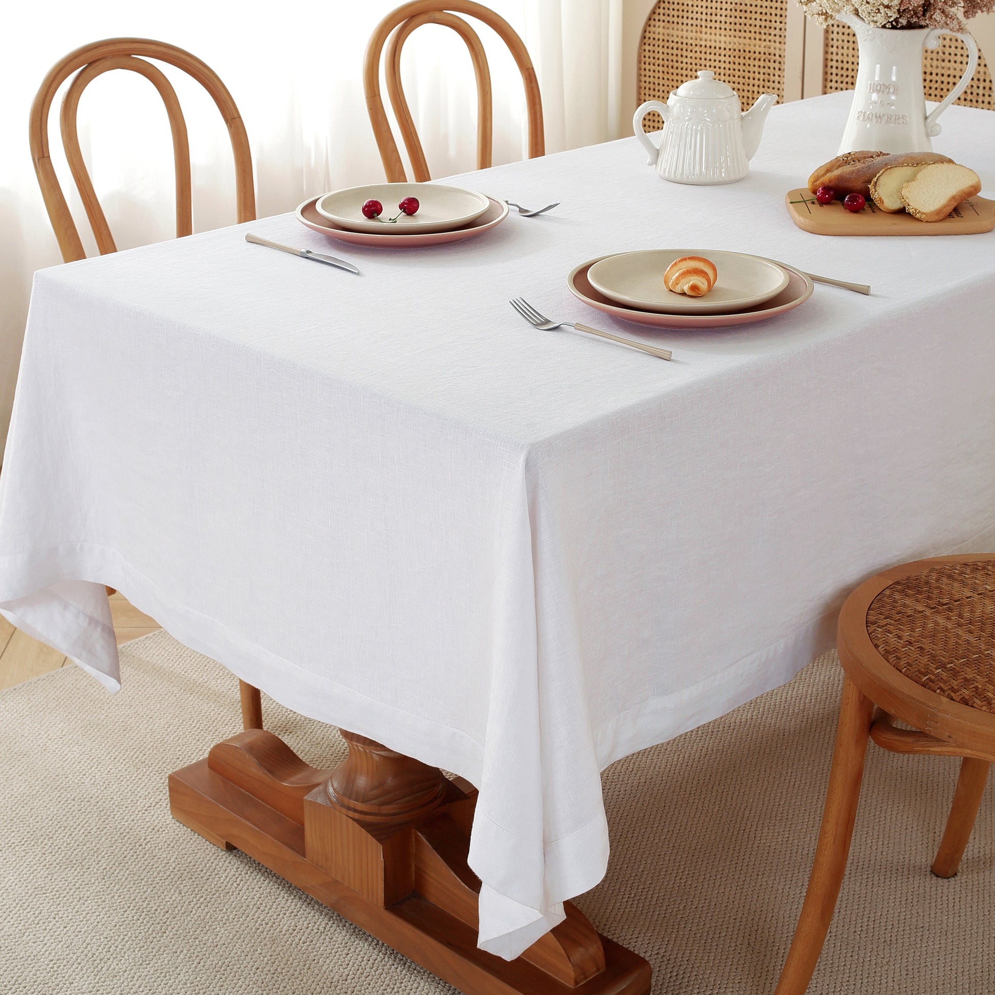 A white tablecloth draped over a wooden dining table, styled with plates, cutlery, a teapot, and bread, with wooden chairs around it.