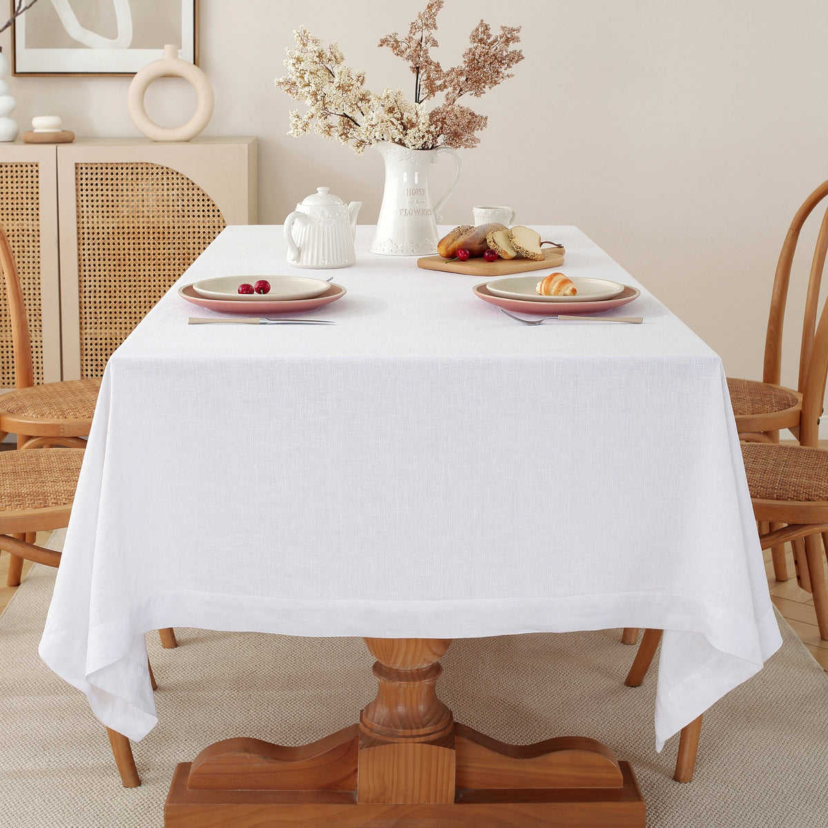 A white tablecloth draped over a wooden dining table, styled with plates, a teapot, and a vase of dried flowers in a warm, neutral-toned room.