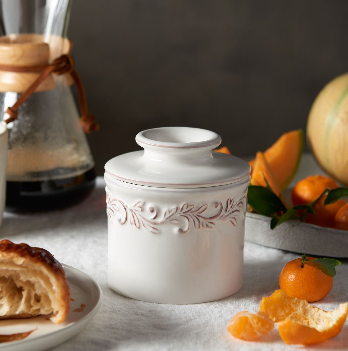 White ceramic jar with decorative patterns on a table with oranges and a coffee filter.