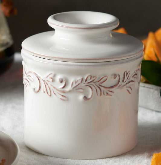 A white ceramic lidded container with embossed floral scrollwork decoration around the middle, shown on a light fabric surface with soft blurred background elements.