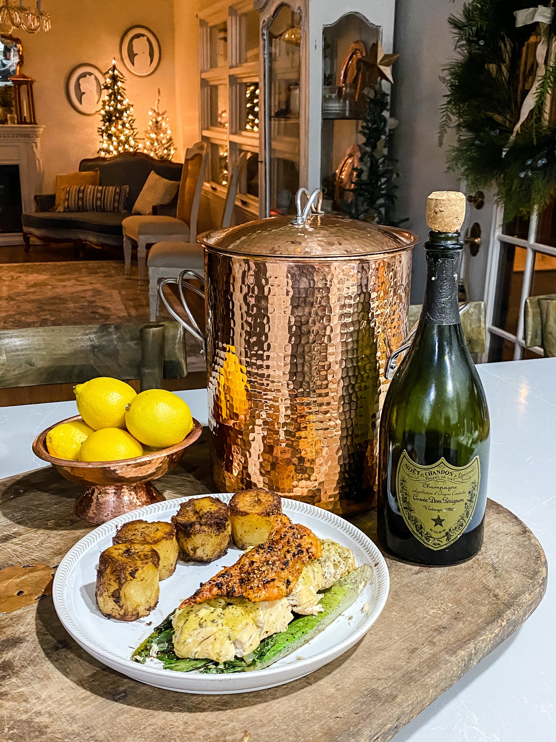 Dinner setup with copper stock pot, seafood dish, lemons, and a bottle of champagne on a wooden board.