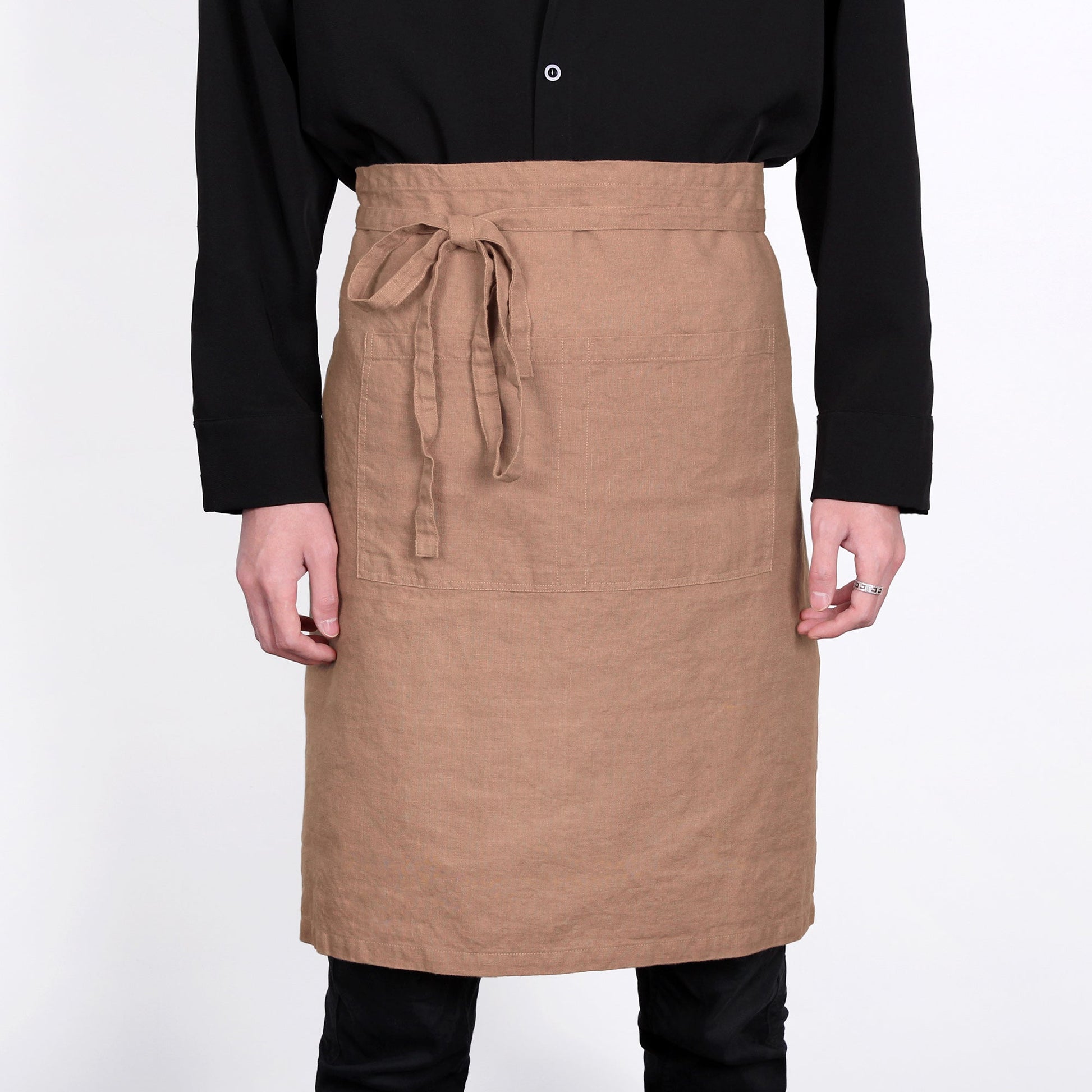 A person wearing a tan linen apron with a tie waist and front pockets, paired with a black long-sleeve shirt and black pants, against a plain white background.