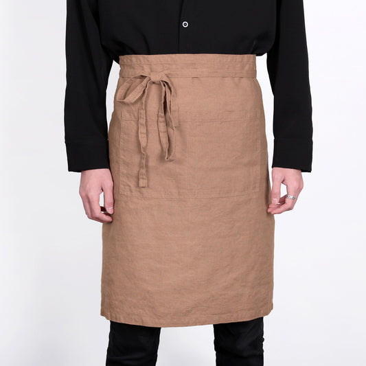 A person wearing a tan linen apron with a tie waist and front pockets, paired with a black long-sleeve shirt and black pants, against a plain white background.