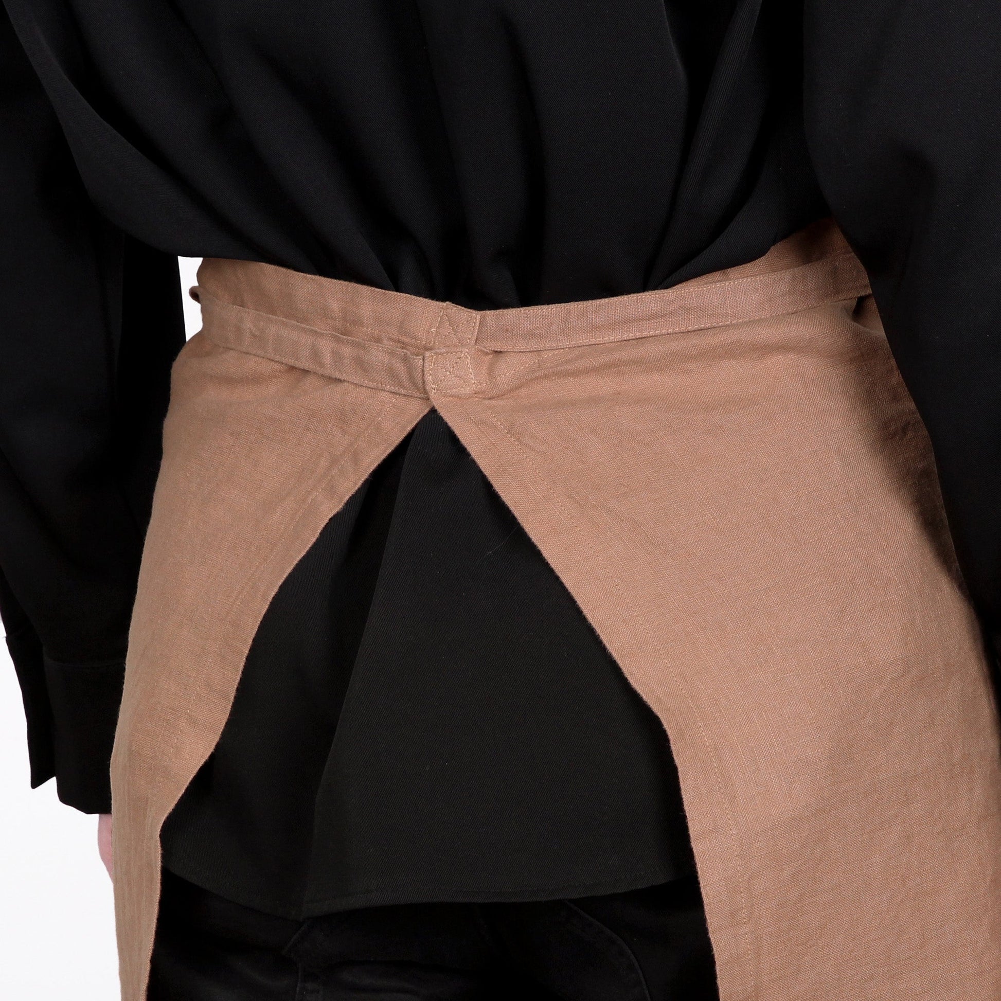 A close-up of a person wearing a light tan linen apron tied around the waist over a black shirt.
